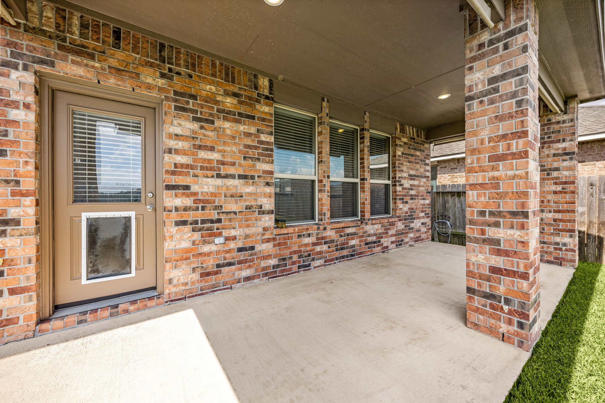 Inviting covered back porch highlighting a full brick accent wall, a triple window bank, and a modern exterior door equipped with a built-in pet door.