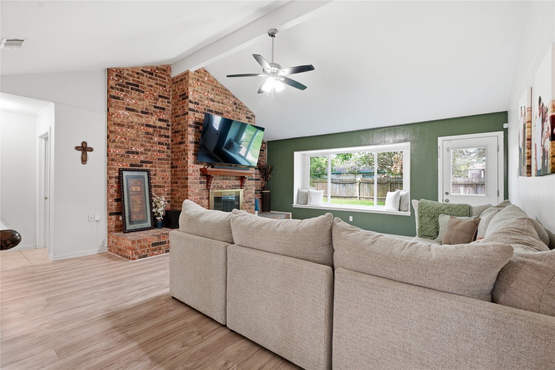 Bright, open living room featuring vaulted ceilings, a brick fireplace, large picture window, and access to the backyard.