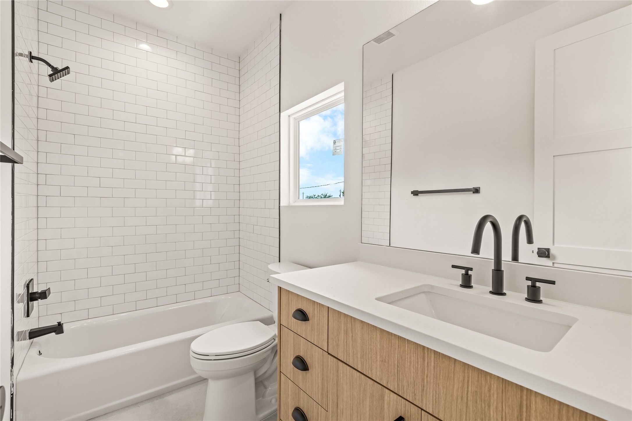 Modern guest bathroom featuring a light wood vanity with a white countertop, matte black hardware, a large frameless mirror, and a classic white subway-tiled tub/shower combination.