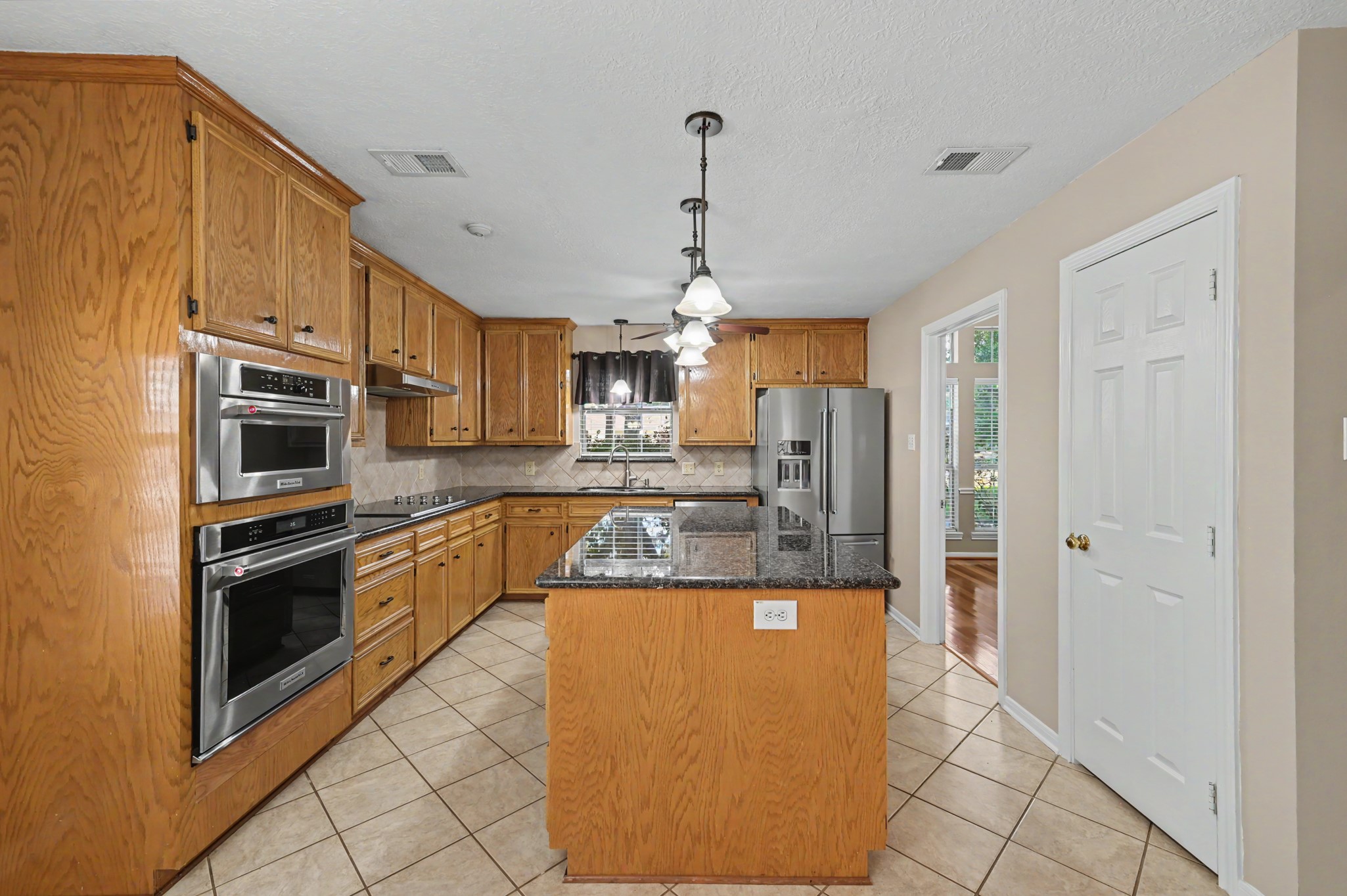 Spacious kitchen with warm wood cabinetry, granite countertops, and sleek stainless steel appliances, featuring a central island and elegant pendant lighting.