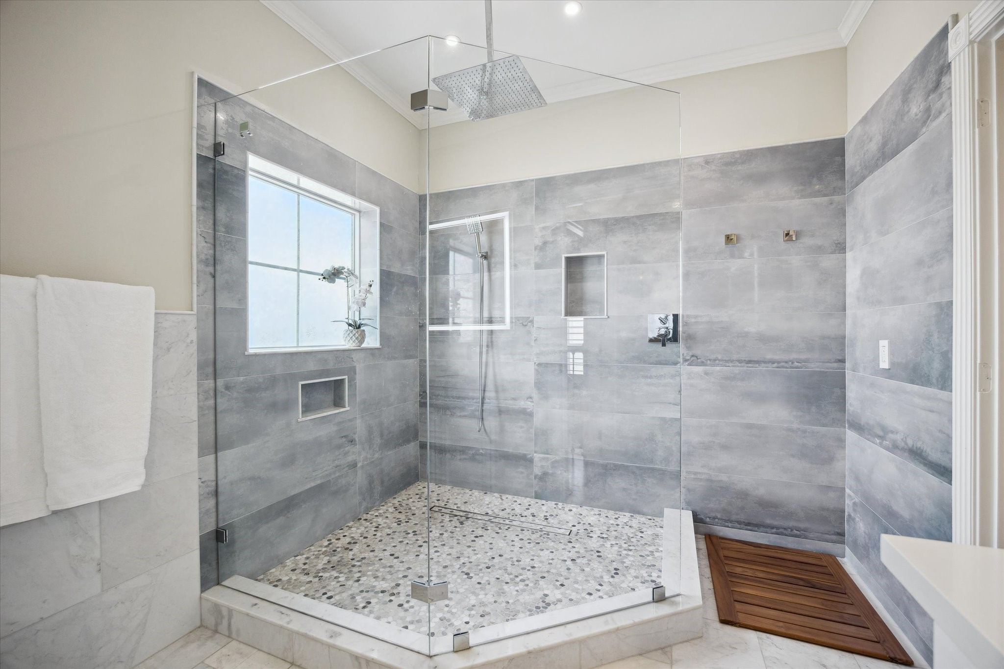 This spacious bathroom features a modern, glass-enclosed shower with elegant gray tiles and a pebble stone floor. It includes built-in niches for storage and a rain shower head for a luxurious experience.