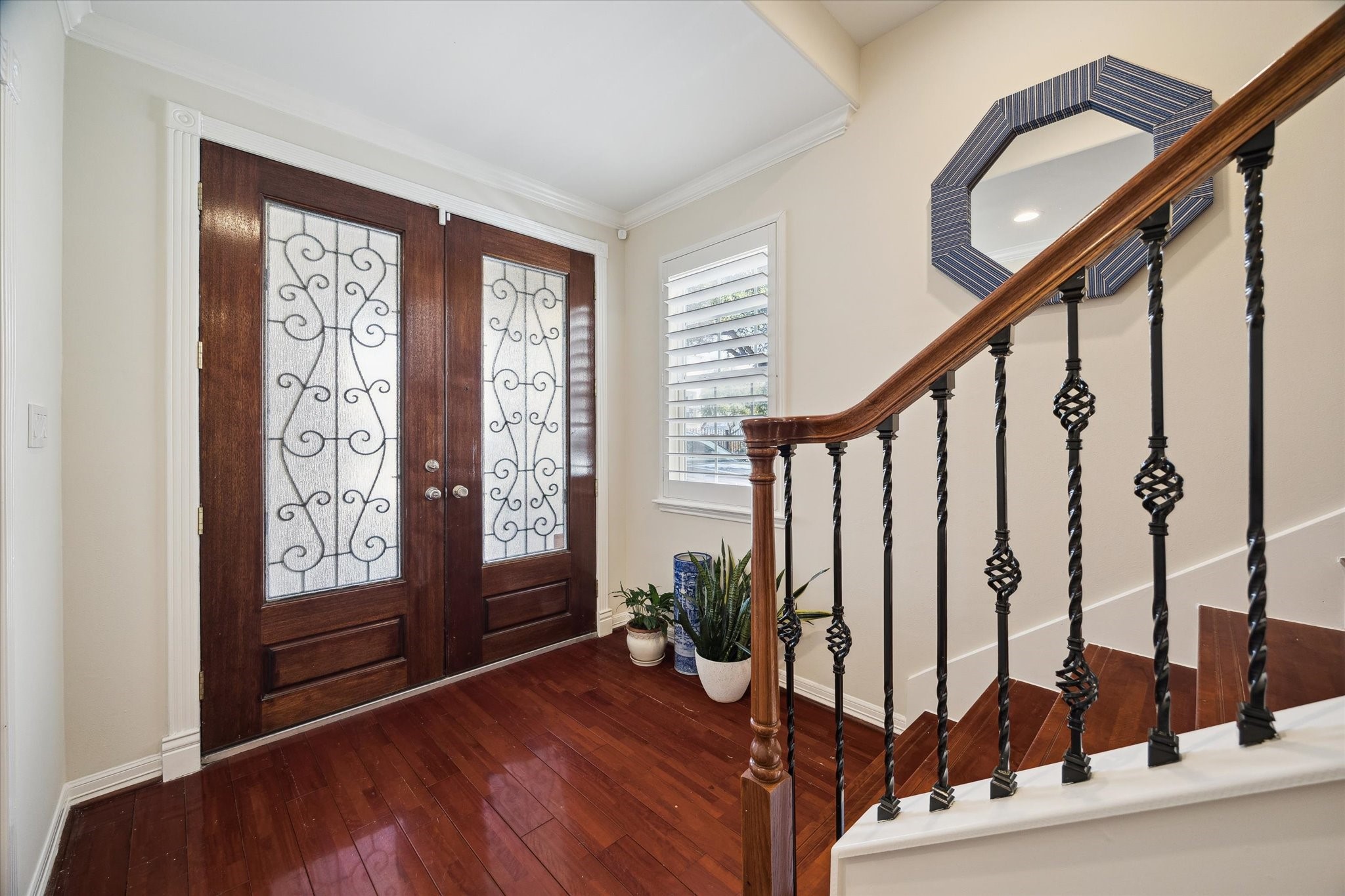 This inviting entryway features elegant double wooden doors with decorative glass and ironwork. It has rich hardwood floors and a stylish staircase with wrought iron balusters. A window with plantation shutters adds natural light.