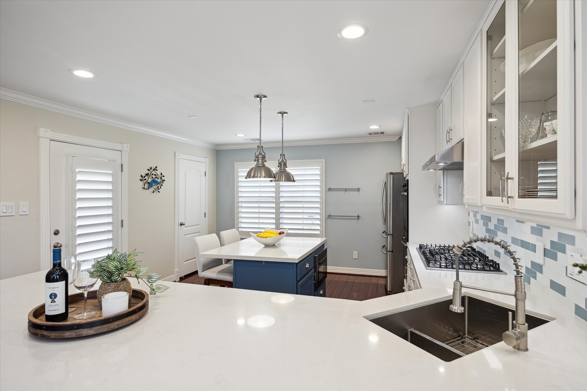 The kitchen features a bright, open layout with white cabinetry, a mosaic backsplash, and a sleek island with bar seating. Stainless steel appliances and ample counter space provide functionality, while pendant lighting and plantation shutters add a stylish touch.