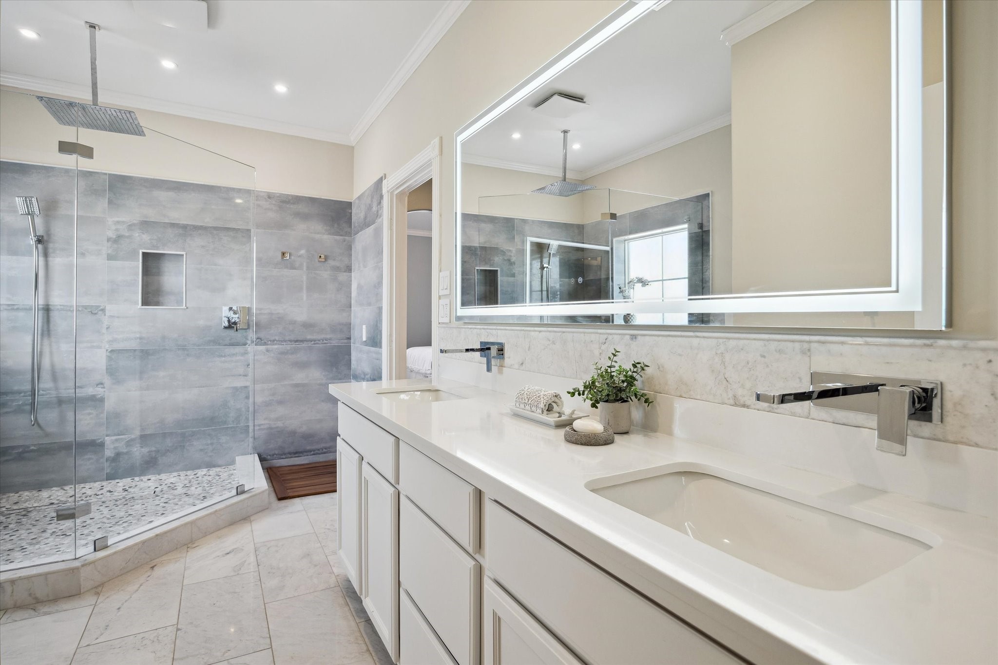 This modern bathroom features a spacious glass-enclosed shower with sleek gray tiles, dual rainfall showerheads, and a pebble stone floor. The double vanity has a marble countertop, contemporary fixtures, and an illuminated mirror, offering a luxurious and functional space.
