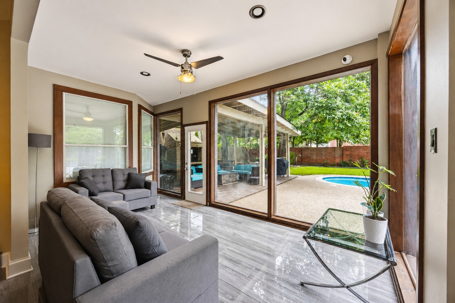 The bright sunroom boasts large wood-framed windows, a ceiling fan, and glossy wood-look tile floors that overlook the pool area.