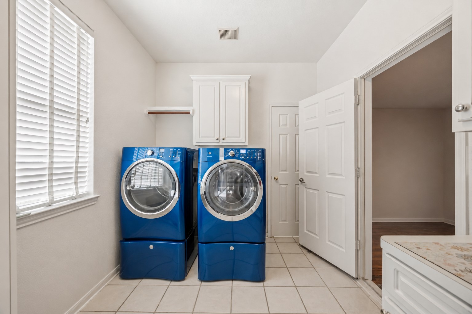 The dedicated laundry room includes white upper cabinetry, a hanging rod, tile floors, and a side window with horizontal blinds.
