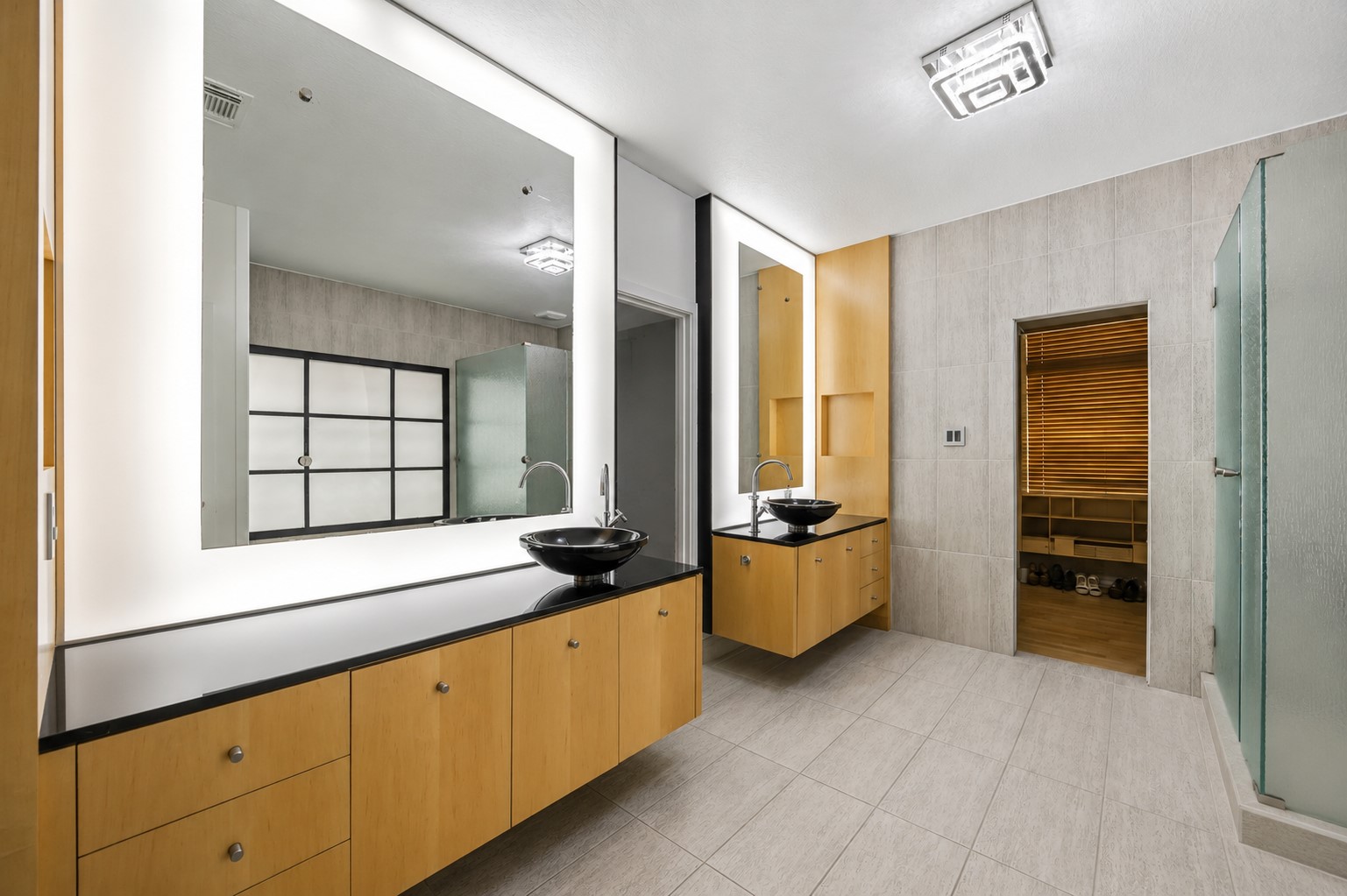 This modern bathroom showcases dual floating vanities with light wood cabinetry, sleek black countertops with vessel sinks, and integrated back-lit mirrors.