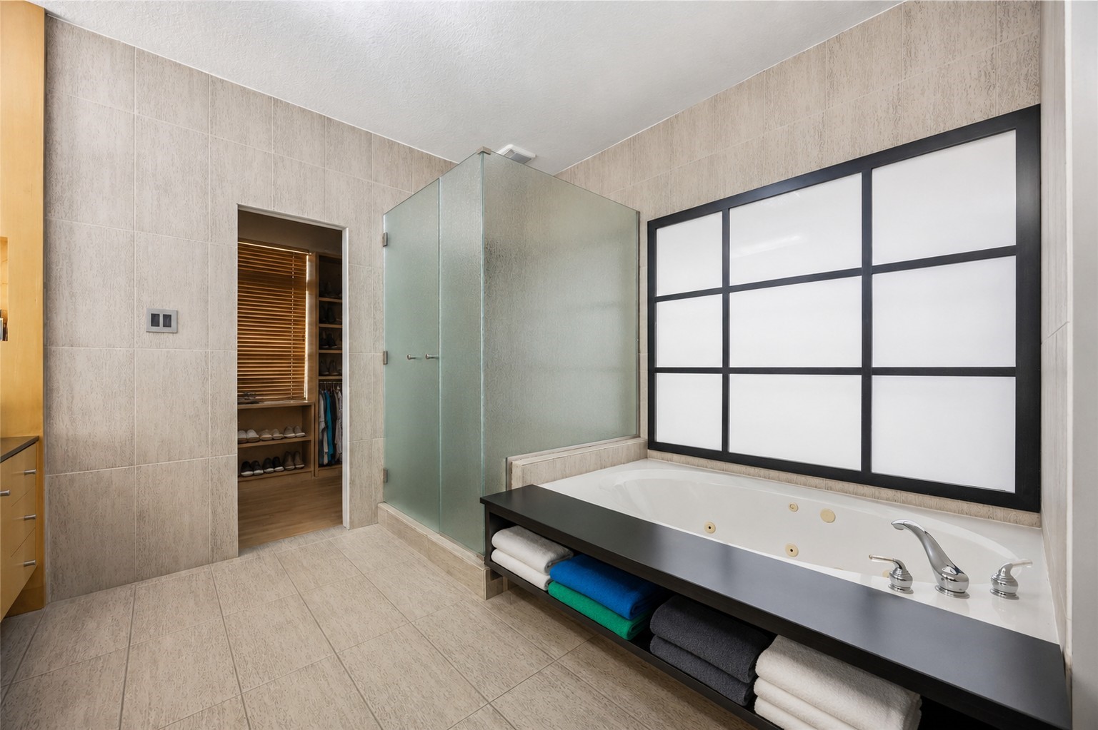 The spa-like bath area includes a glass-enclosed walk-in shower, tile flooring, and a deep soaking tub set against a large grid-patterned privacy window.