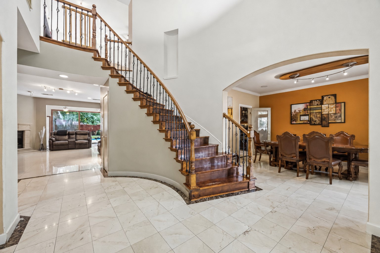 The bright entryway showcases polished tile flooring with a dark border inlay, a sweeping wooden staircase with wrought-iron spindles, and an arched transition into the adjacent dining area.