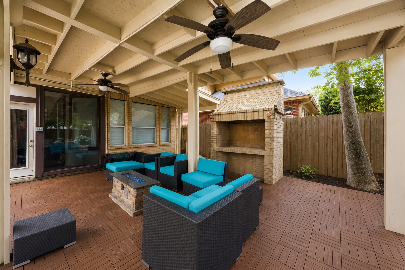 The covered veranda boasts a rustic exposed-joist ceiling with cooling fans, patterned interlocking deck tiles, and a large built-in brick outdoor fireplace kitchen.