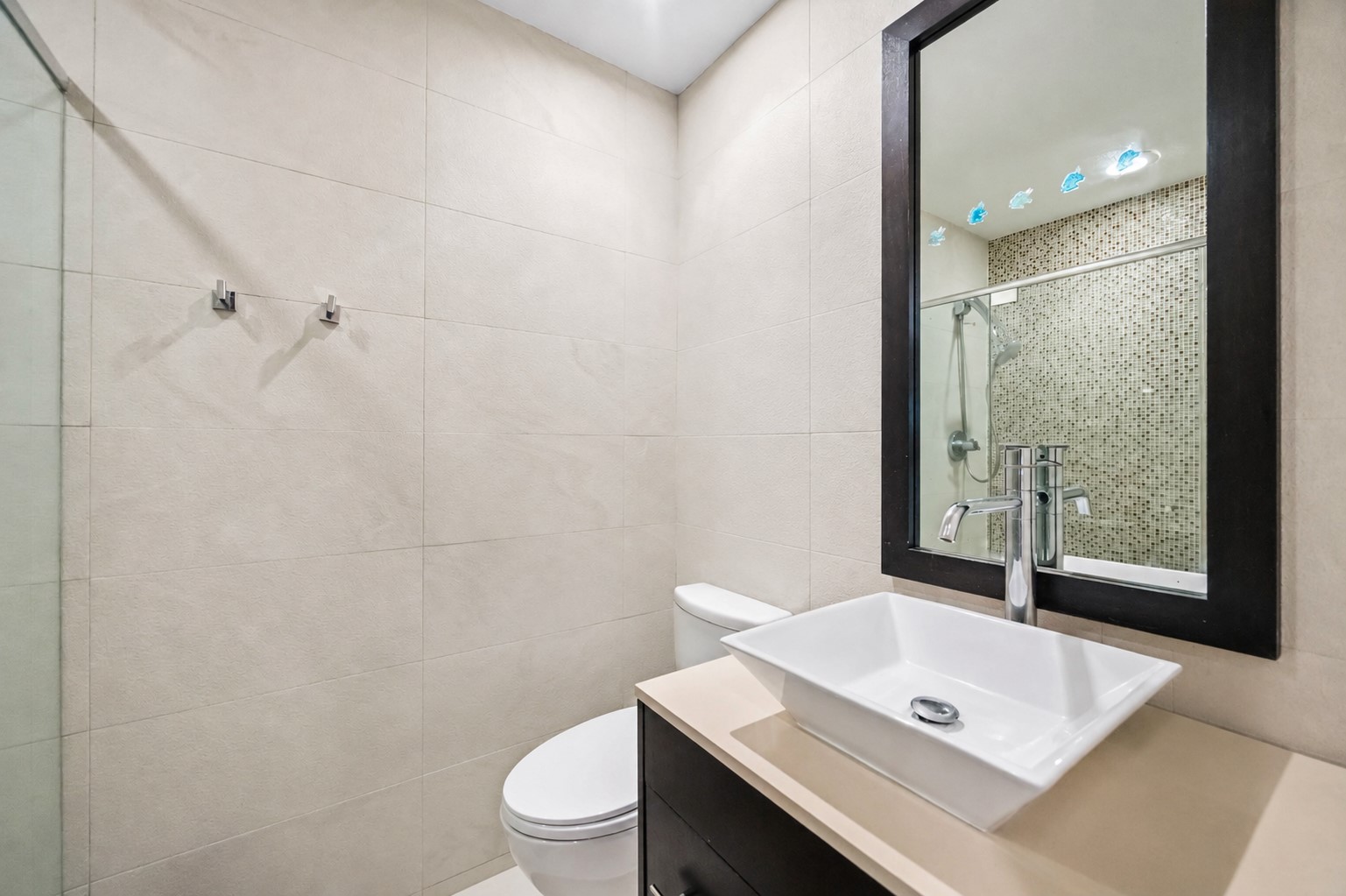 This bathroom spaBathroom with neutral tile, dark floating vanity, white countertop, vessel sink, and framed mirror.ce features premium neutral wall tiling, a dark floating vanity cabinet with a sleek white countertop, and a vessel sink beneath a dark-framed mirror.