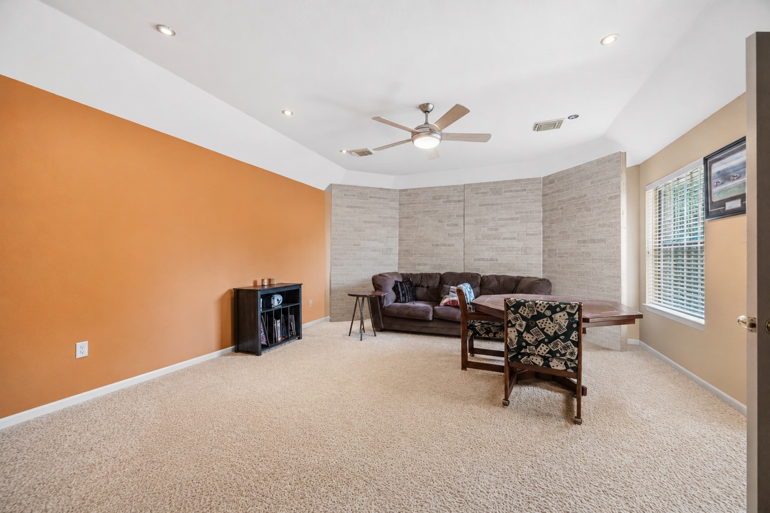 This expansive upstairs bedroom features plush beige carpeting, a central ceiling fan, an orange accent wall, and a unique textured brick-look focal wall.