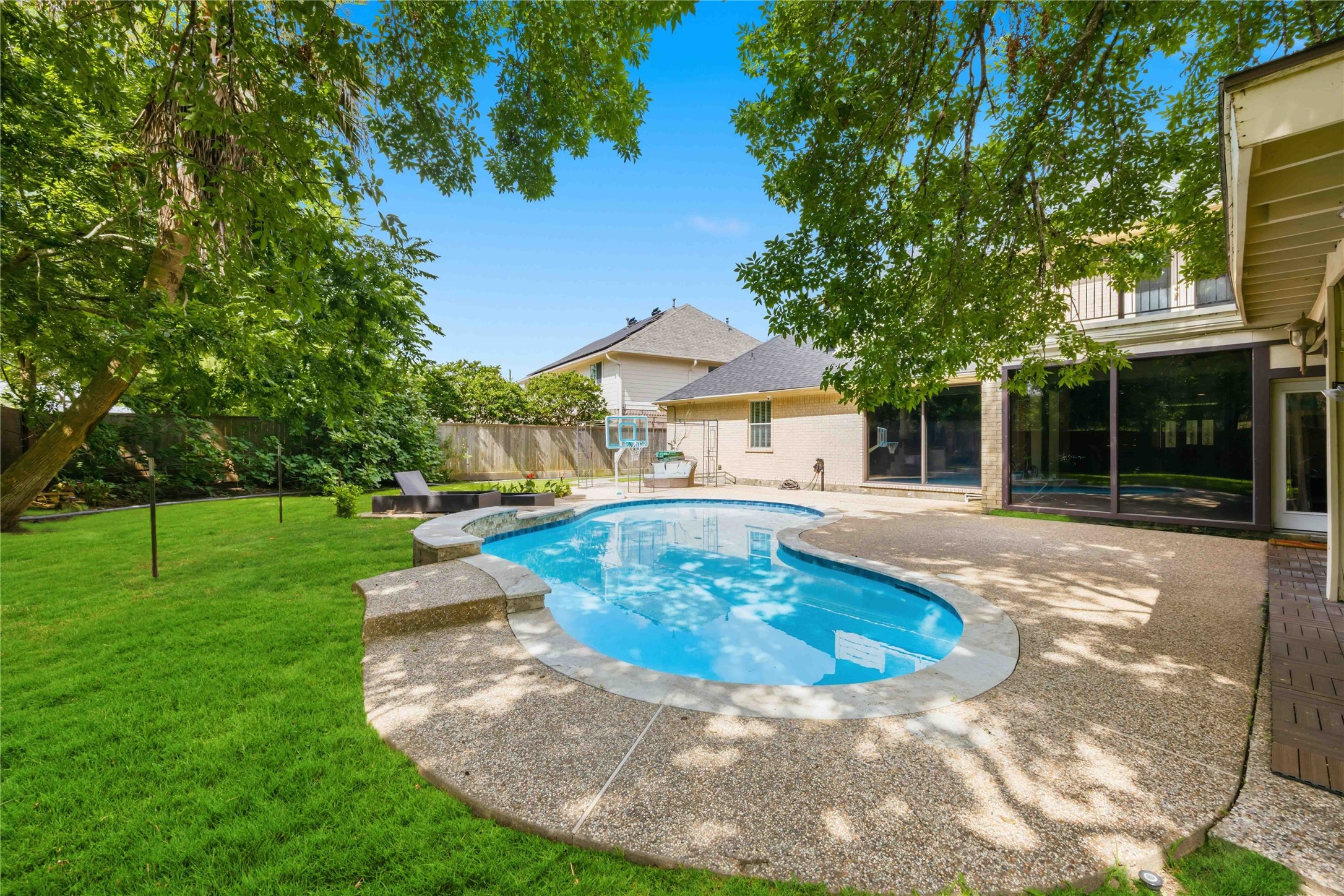 A wide lush lawn borders the custom swimming pool, bordered by mature trees and privacy wood fencing.