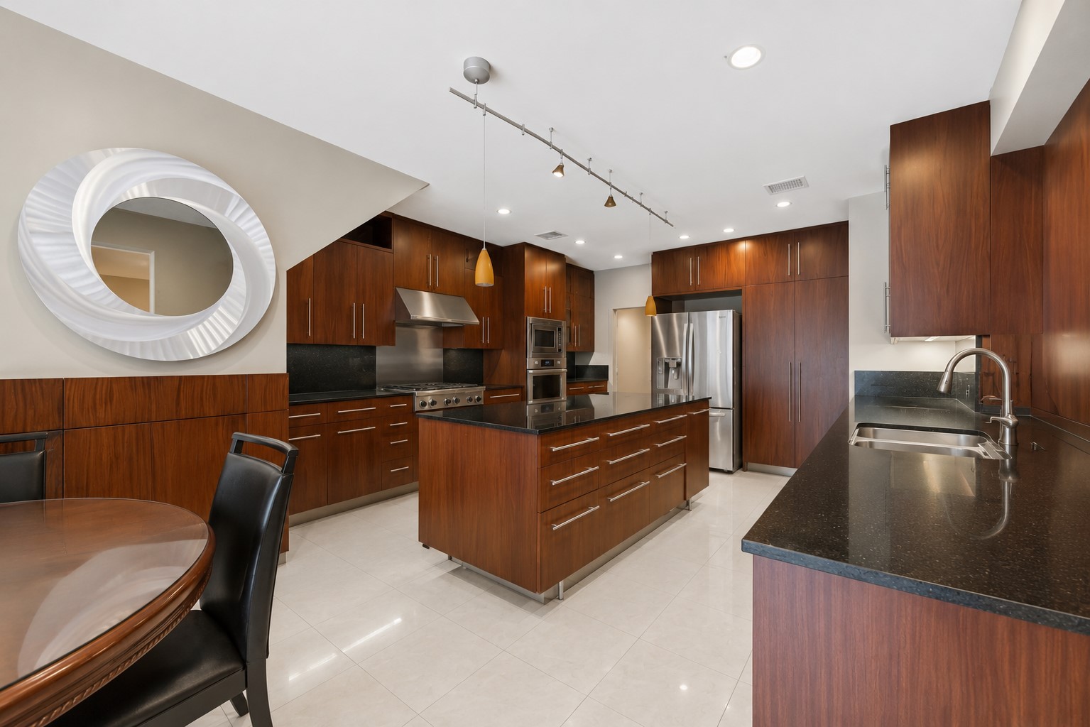 The contemporary kitchen showcases rich wood cabinetry, dark granite countertops, and an expansive central island lit by track lighting.
