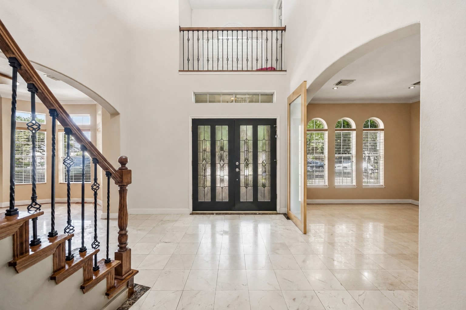 A soaring two-story foyer highlights a dramatic black double-door entry with decorative glass panels, an overhead balcony railing, and large multi-paned windows that fill the space with light.