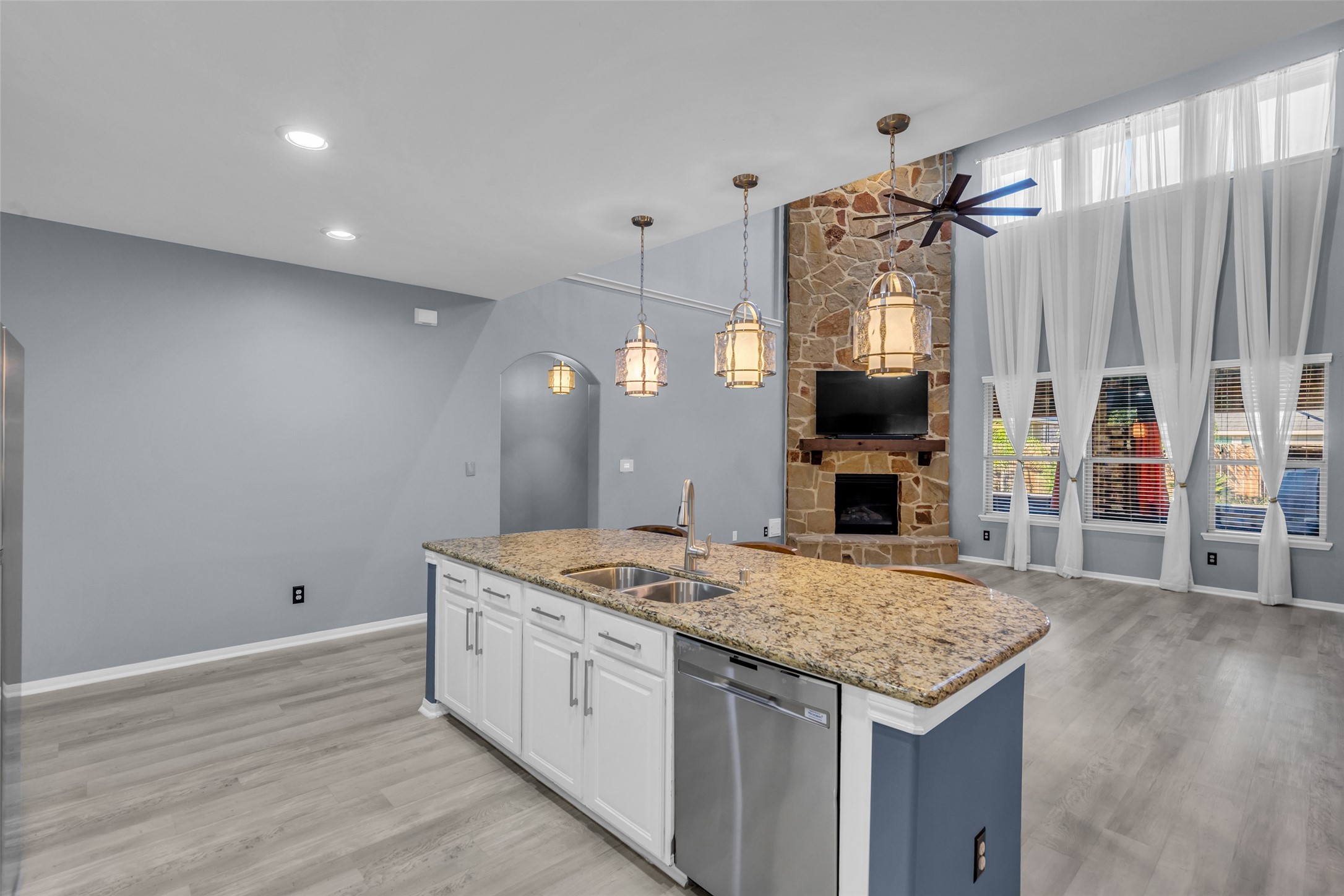 Spacious living area featuring a stunning stone fireplace, high ceilings with elegant drapery, and a modern kitchen island with granite countertops and pendant lighting.
