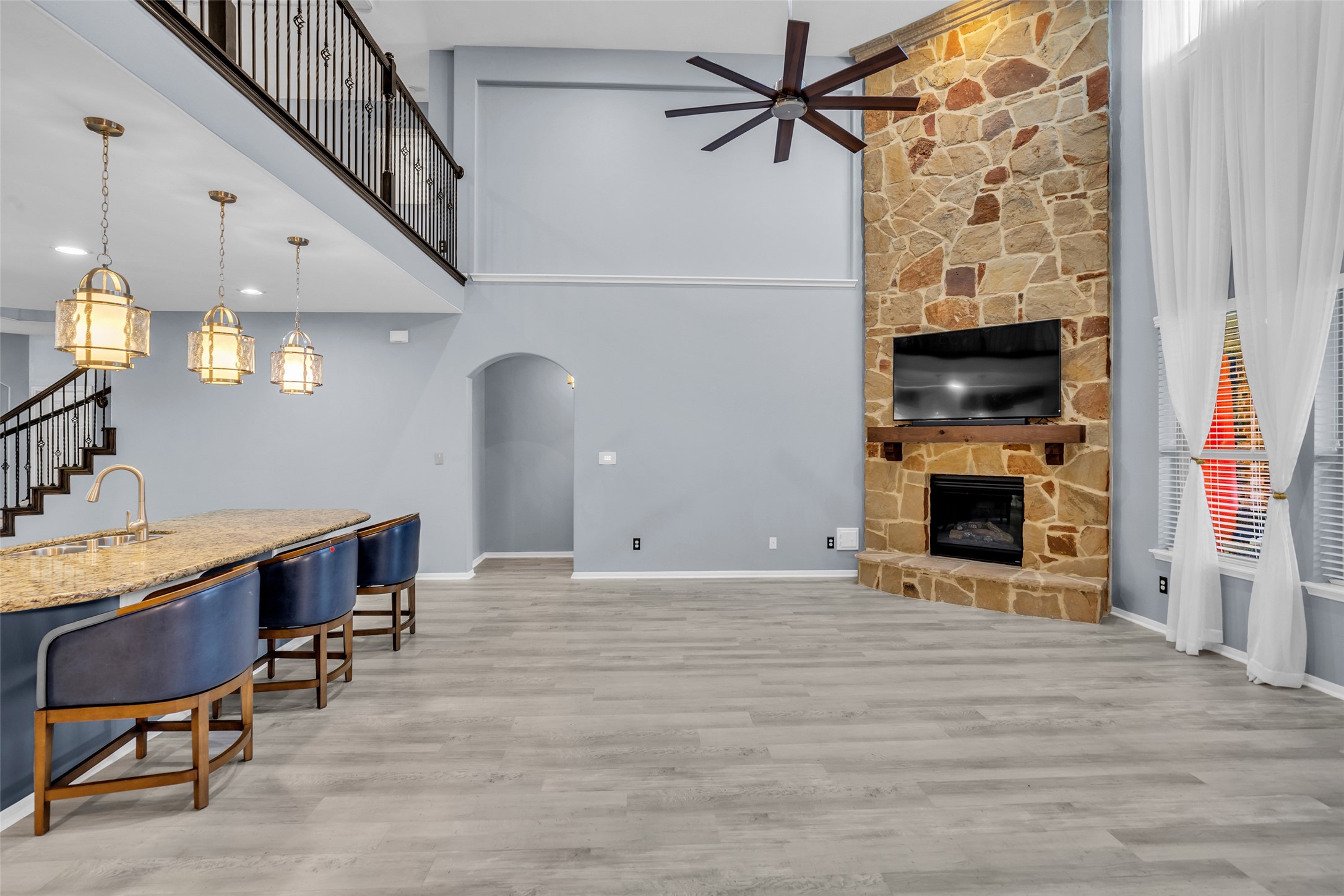 Spacious living area with a stunning stone fireplace, modern ceiling fan, and large windows that fill the room with natural light. The open layout includes a sleek kitchen island with elegant pendant lighting and bar seating, perfect for entertaining.