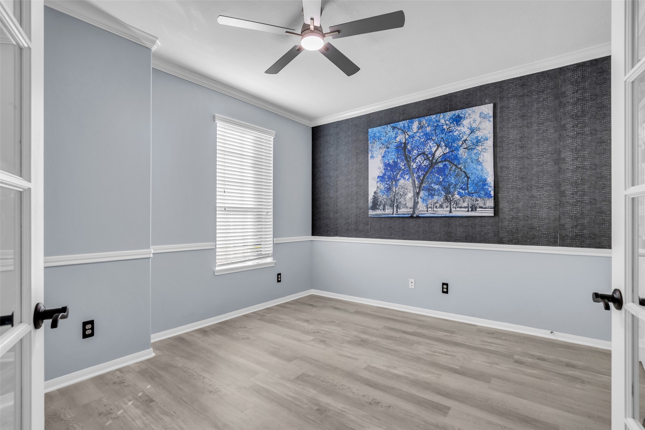 Bright and inviting office featuring elegant gray walls with white trim, stylish wood flooring, and a contemporary ceiling fan. A striking piece of art adds a pop of color, perfect for a home office or cozy retreat.