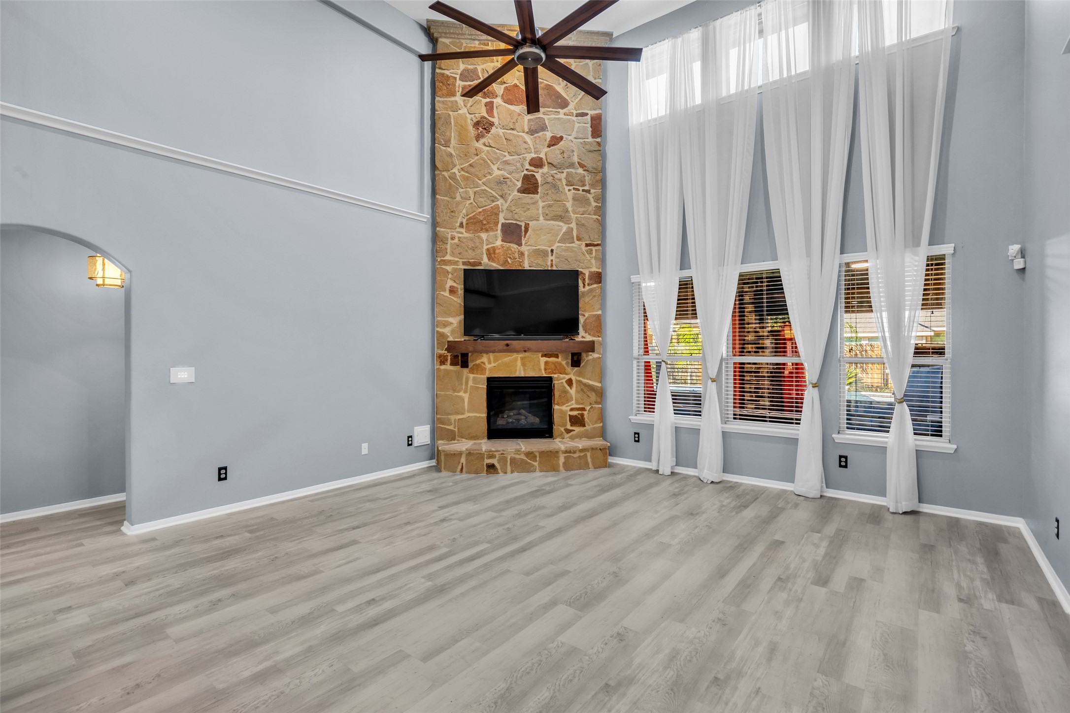 Bright and spacious living room featuring soaring ceilings, a stunning stone fireplace, and large windows adorned with elegant sheer curtains, creating a warm and inviting atmosphere.