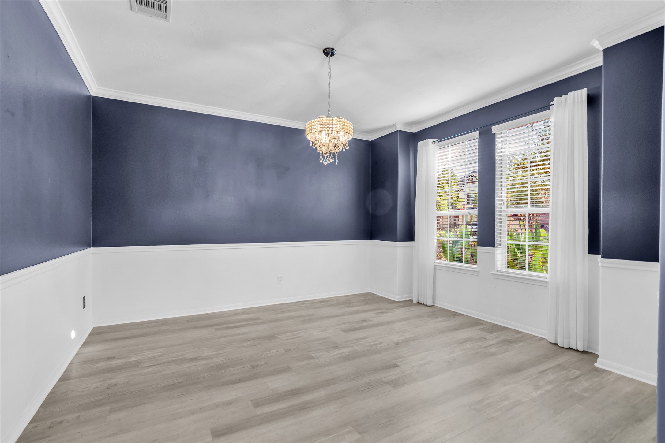 Elegant formal dining room featuring rich blue walls with white wainscoting, accented by a classic chandelier and large windows that fill the space with natural light.