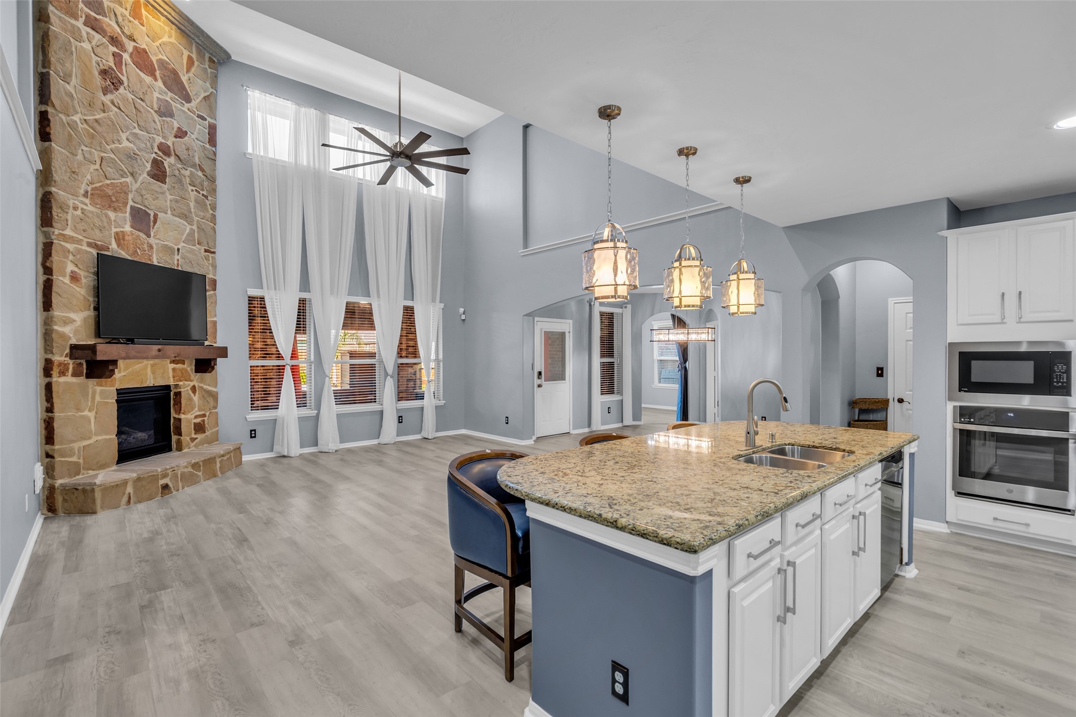 Spacious open-concept living area featuring a stunning stone fireplace, high ceilings with a modern fan, and a gourmet kitchen with granite countertops and elegant pendant lighting.