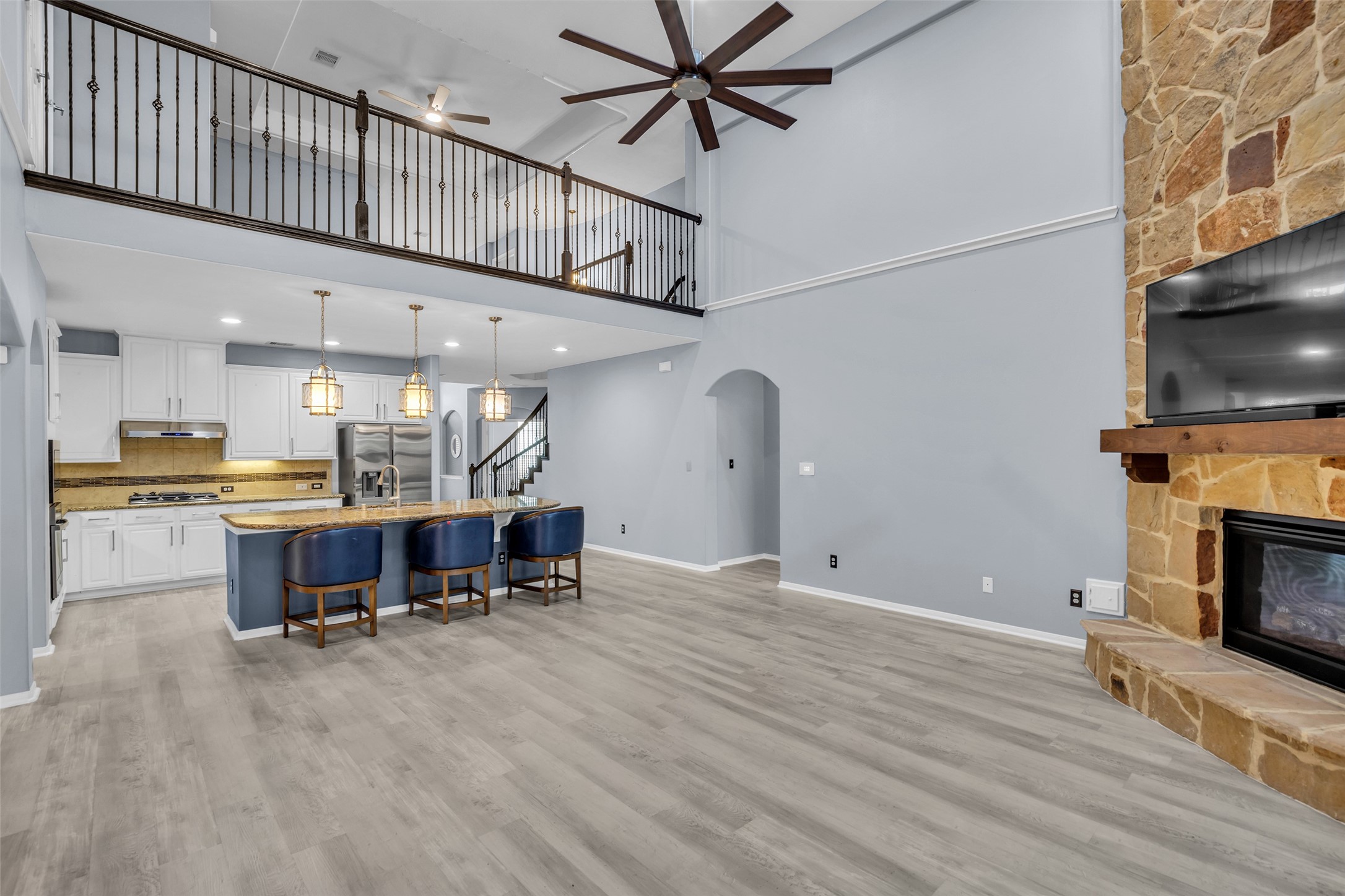 Spacious open-concept living area featuring a vaulted ceiling, elegant stone fireplace, and modern kitchen with an island bar. The loft-style railing adds a touch of sophistication, complemented by sleek wood flooring and ample natural light.