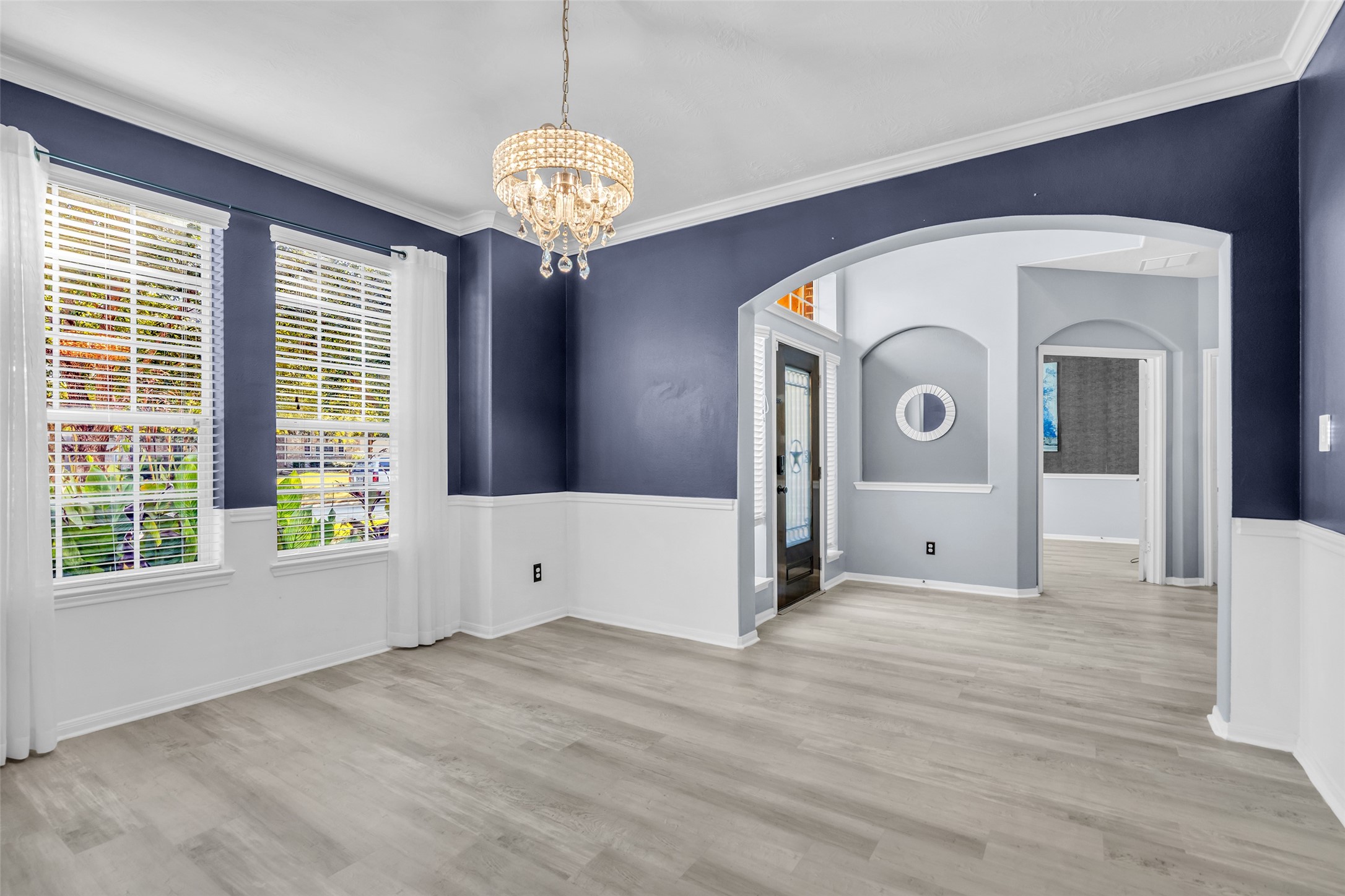 Elegant formal dining area featuring bold navy walls, white wainscoting, and a sparkling chandelier, complemented by abundant natural light from large windows and stylish archways leading to adjacent rooms.