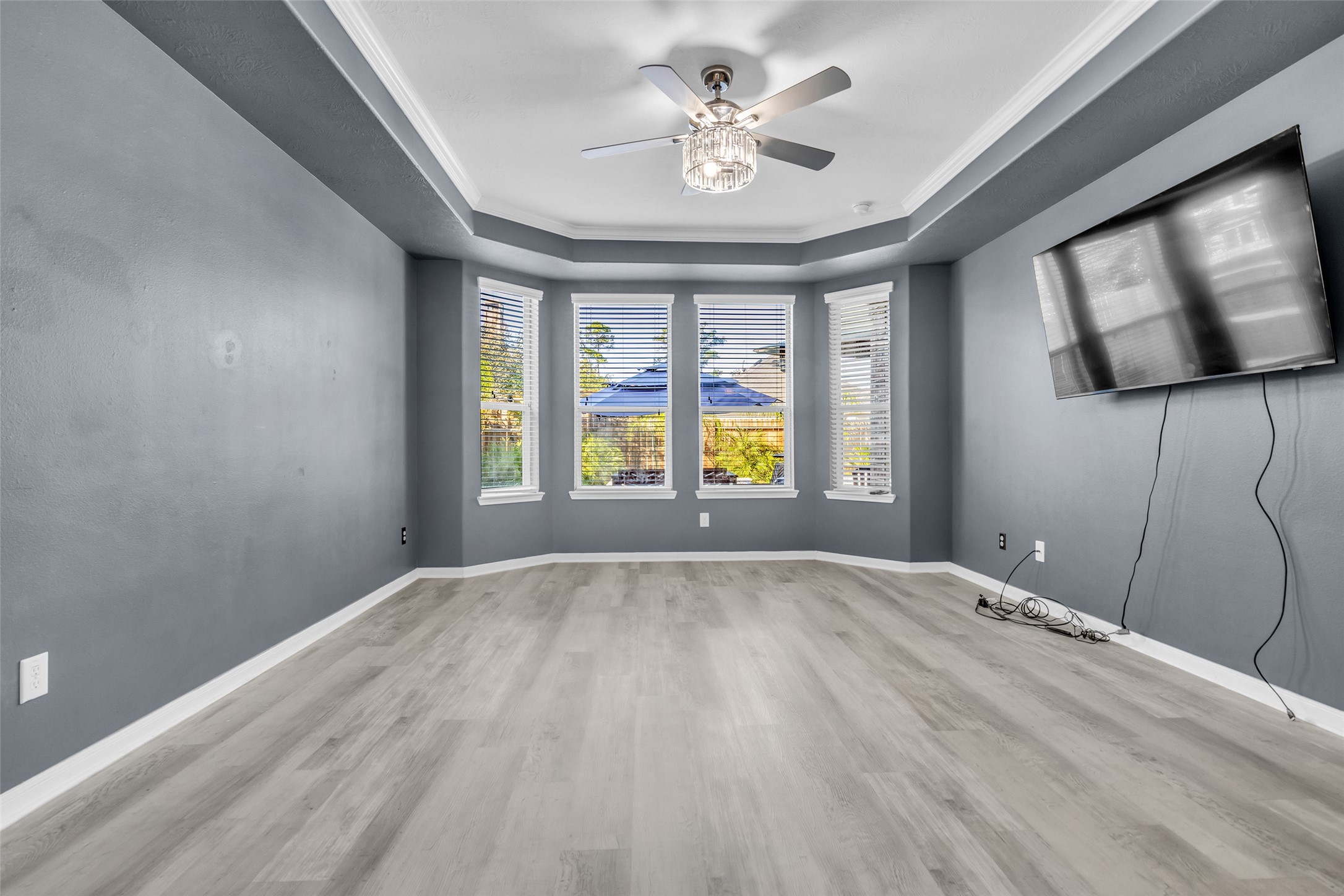 Spacious primary bedroom featuring elegant tray ceiling, chic gray walls, and abundant natural light from large bay windows, complemented by modern flooring and a ceiling fan.
