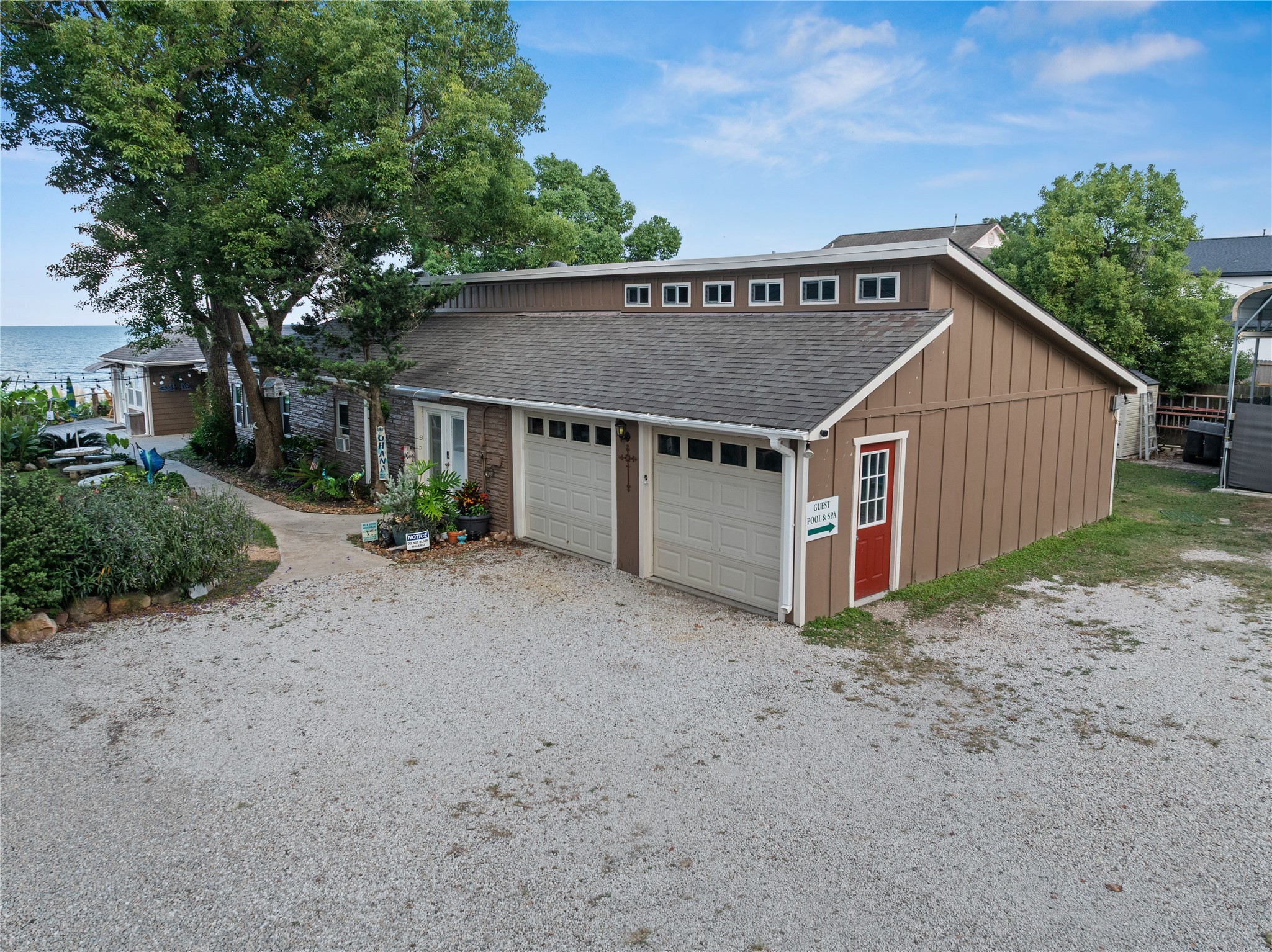 This photo showcases the charming waterfront Ohana House with a rustic exterior, featuring a two-car garage and mature trees. A gravel driveway leads to the property, which offers scenic water views and a cozy atmosphere. Perfect for those seeking a peaceful retreat.