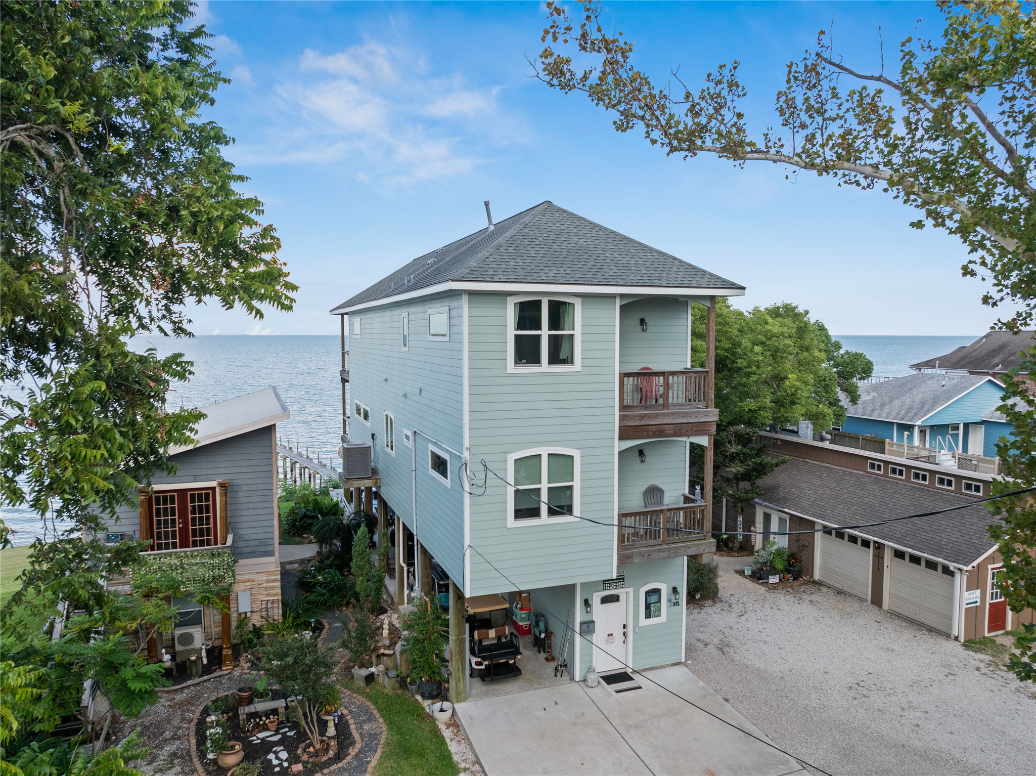 This photo features the charming, elevated coastal home with two stories, featuring a light blue exterior and two balconies with waterfront views. It's nestled among trees with a small landscaped garden and offers bay side living with direct access to the waterfront.