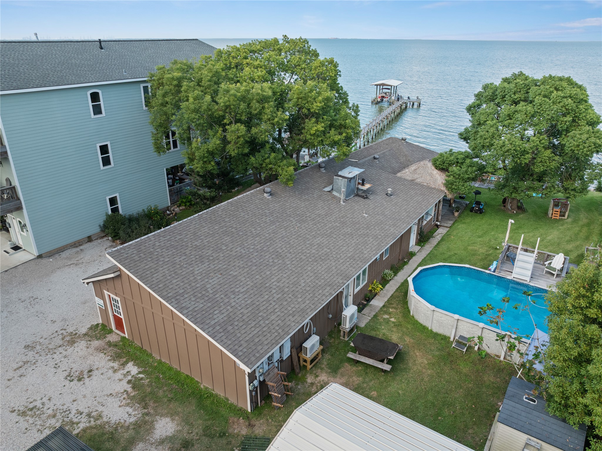 This photo features a spacious one-story Ohana House and Bay Room with a large backyard, a wooden dock extending into the water. Surrounded by greenery, it offers serene views and direct access to the water, perfect for those seeking a peaceful retreat.