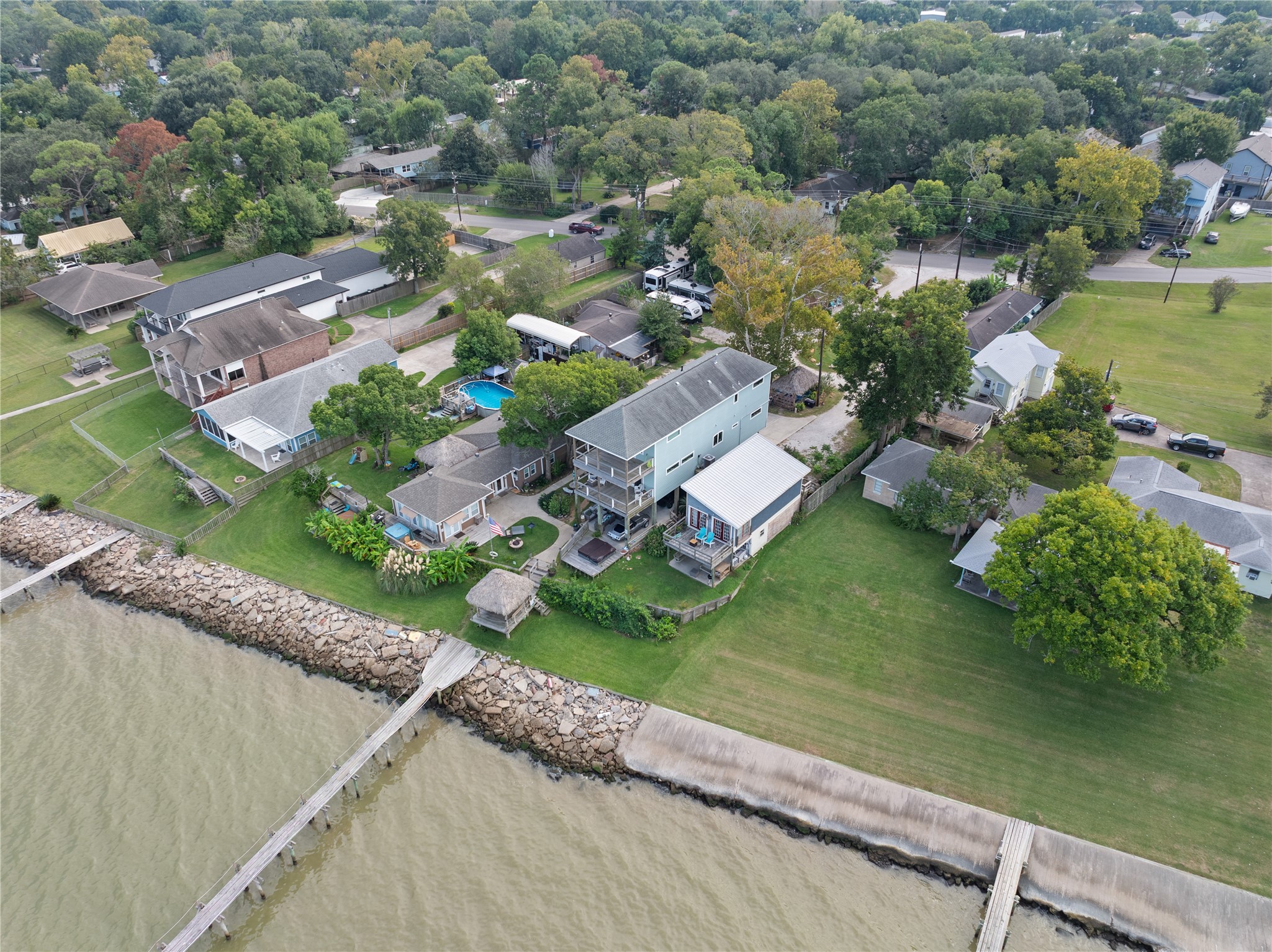 This aerial view showcases the charming waterfront property with a spacious backyard and private pier. The neighborhood is lush with trees, offering a serene, natural setting. Nearby homes are well-maintained, and the area provides a sense of community and privacy.