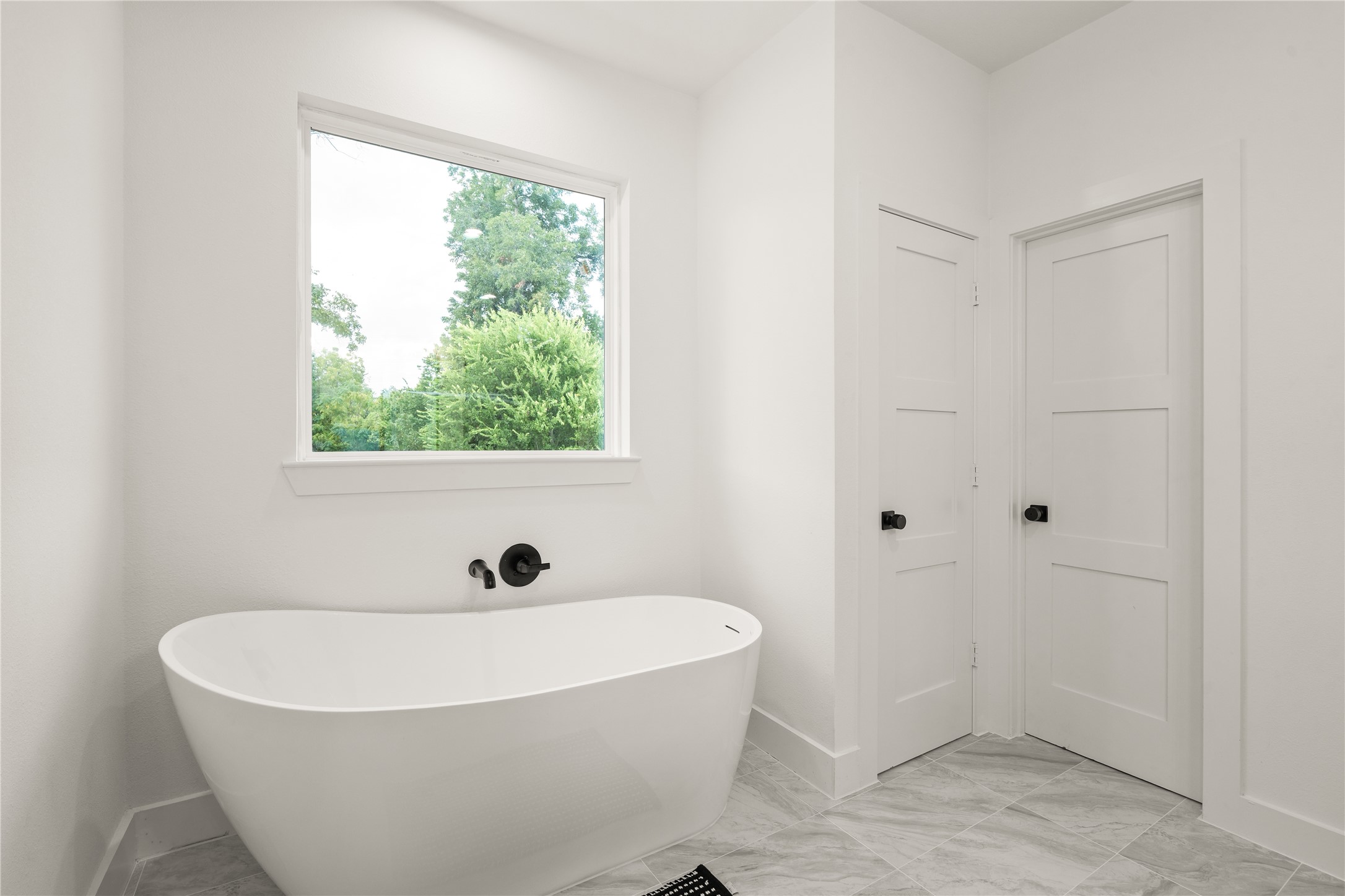 A sculptural freestanding soaking tub sits beneath a picture window framing mature treetops, paired with a
wall-mounted matte-black tub filler for a serene, spa-like moment at the end of the day.
