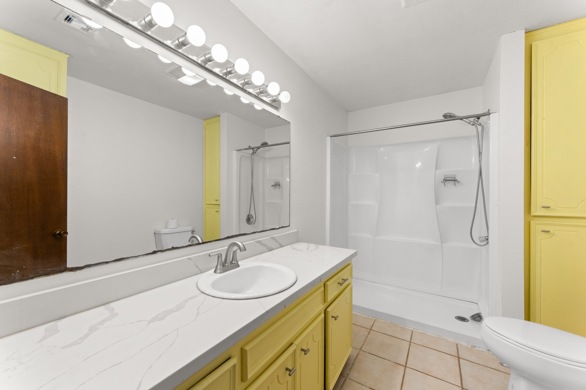 Spacious bathroom featuring a long vanity with marble-look countertops, a large wall mirror, cheerful yellow cabinetry, additional built-in storage, and a walk-in shower with dual showerheads.