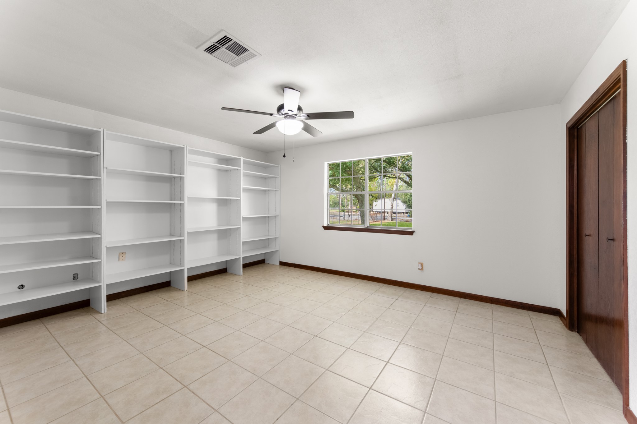 Spacious primary bedroom featuring a wall of removable shelving, tile flooring, and a large window overlooking the property grounds.
