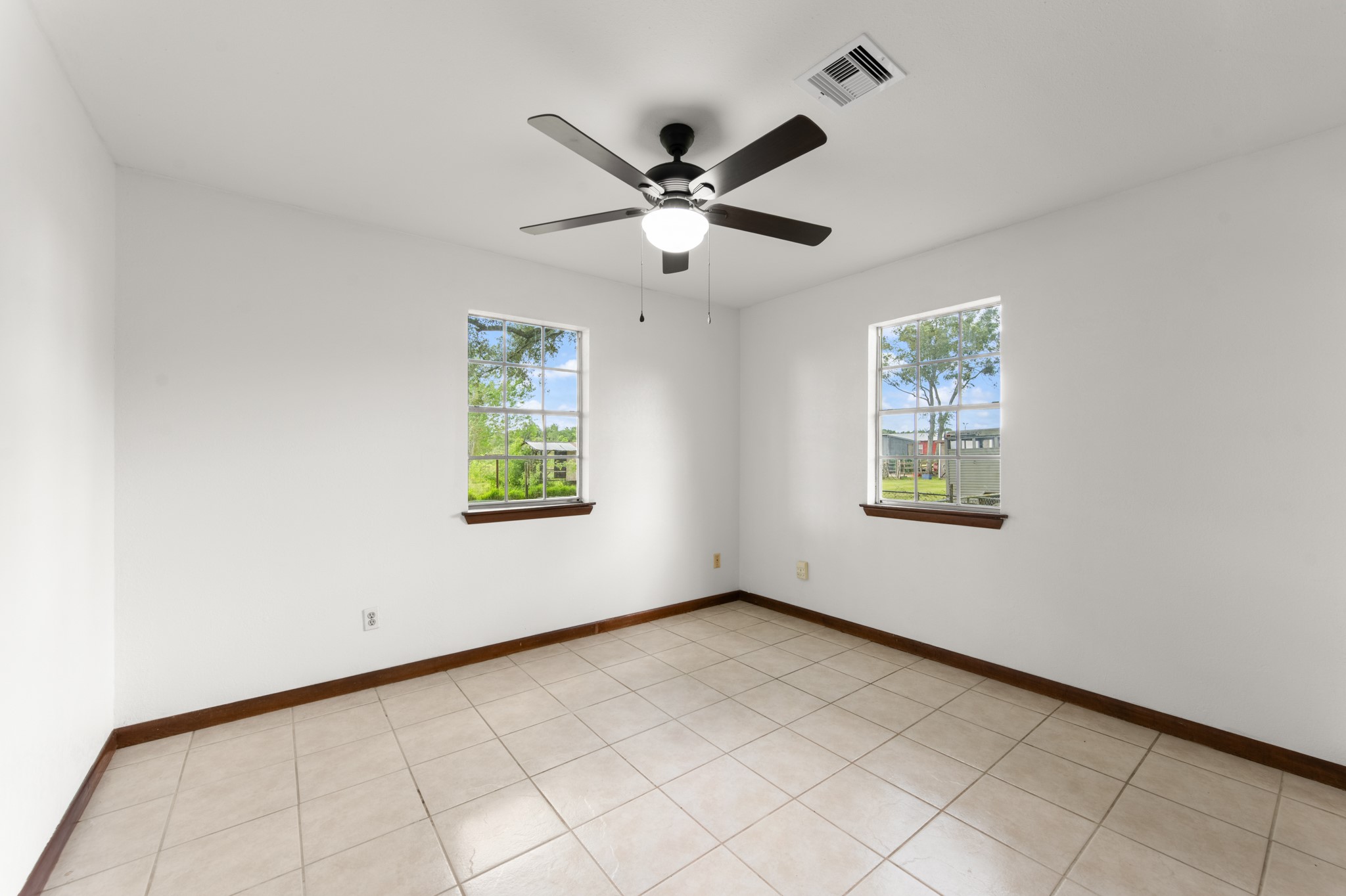 Bright bedroom with two windows overlooking the outdoor surroundings, neutral finishes, tile flooring, and a ceiling fan.