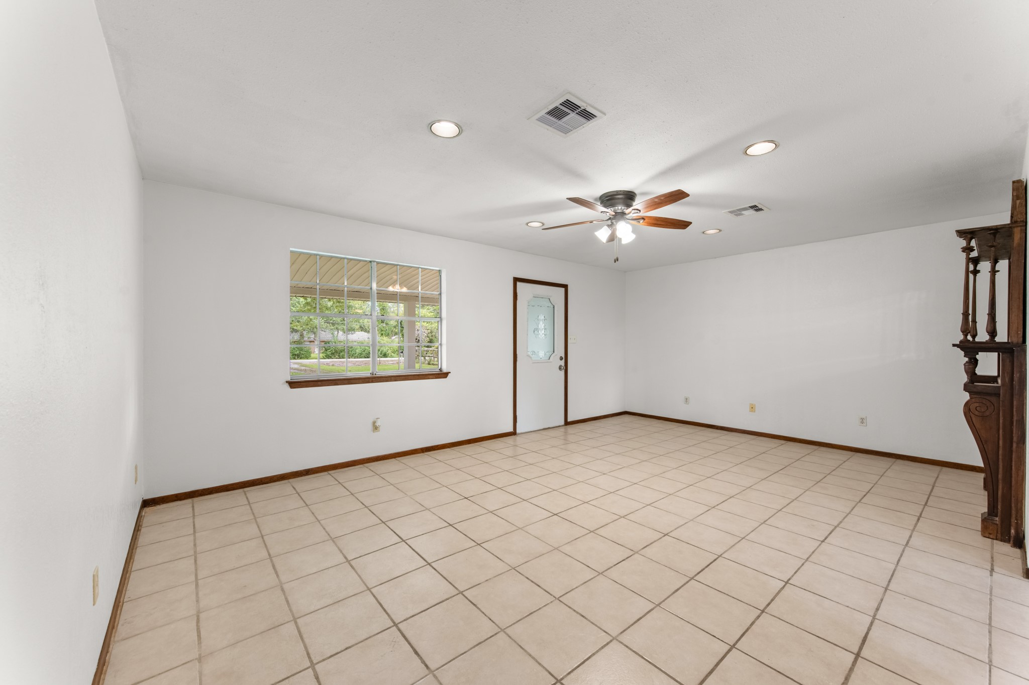 Generously sized living room with neutral walls, easy-care tile flooring, and a ceiling fan. The large window offers views of the covered front porch and mature trees.