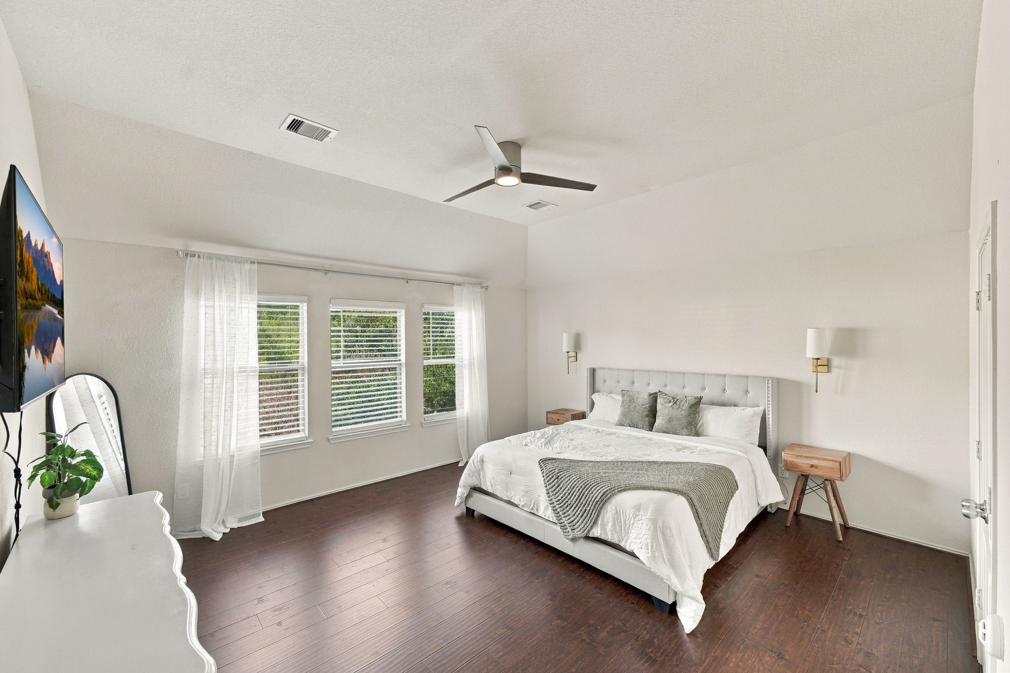 Spacious and bright primary bedroom featuring rich hardwood floors, a large bed with plush bedding, and ample natural light streaming through multiple windows.