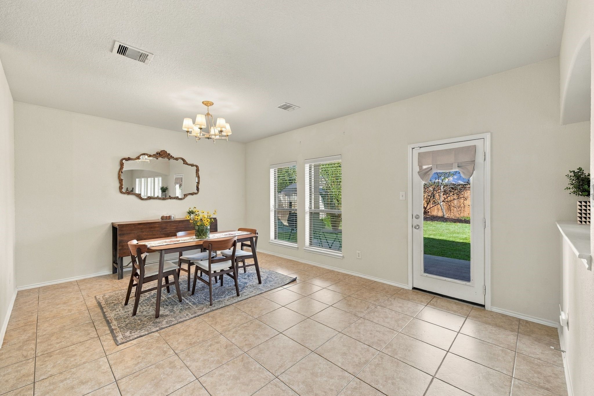 Bright and inviting dining area with tile flooring, a charming chandelier, and double windows allowing natural light to flood the space. The room features a stylish dining set and direct access to a lush backyard through a glass door.