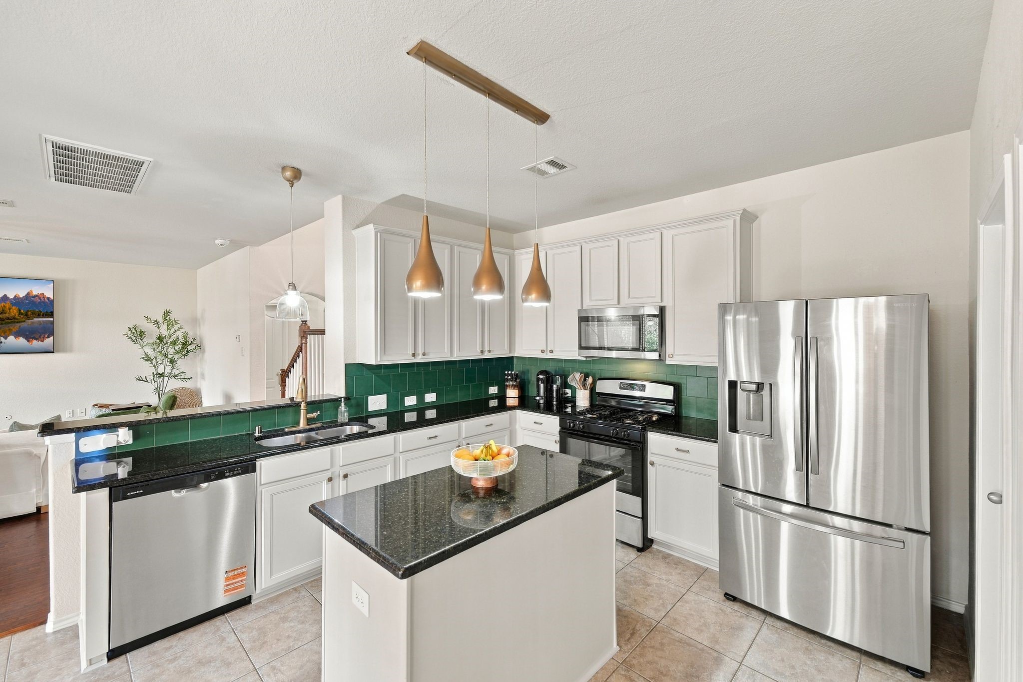 Modern kitchen with sleek stainless steel appliances, a central island with dark granite countertops, and stylish pendant lighting. The bright space features white cabinetry and a deep green tile backsplash, adding a touch of elegance.