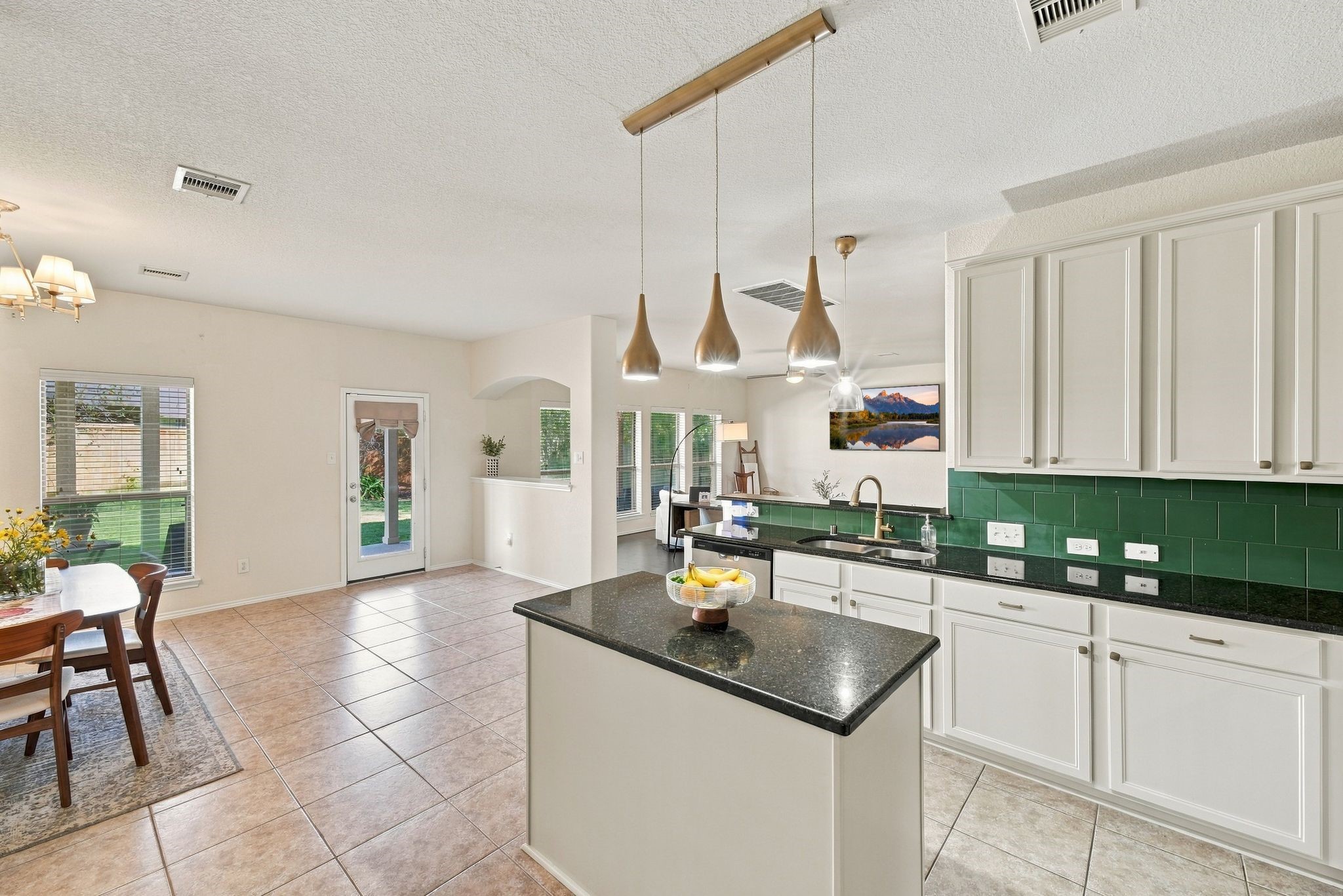 Spacious kitchen featuring sleek white cabinetry, a dark granite countertop island with pendant lighting, and a vibrant green backsplash. The open layout flows seamlessly into a bright dining area with large windows and a view of the backyard.