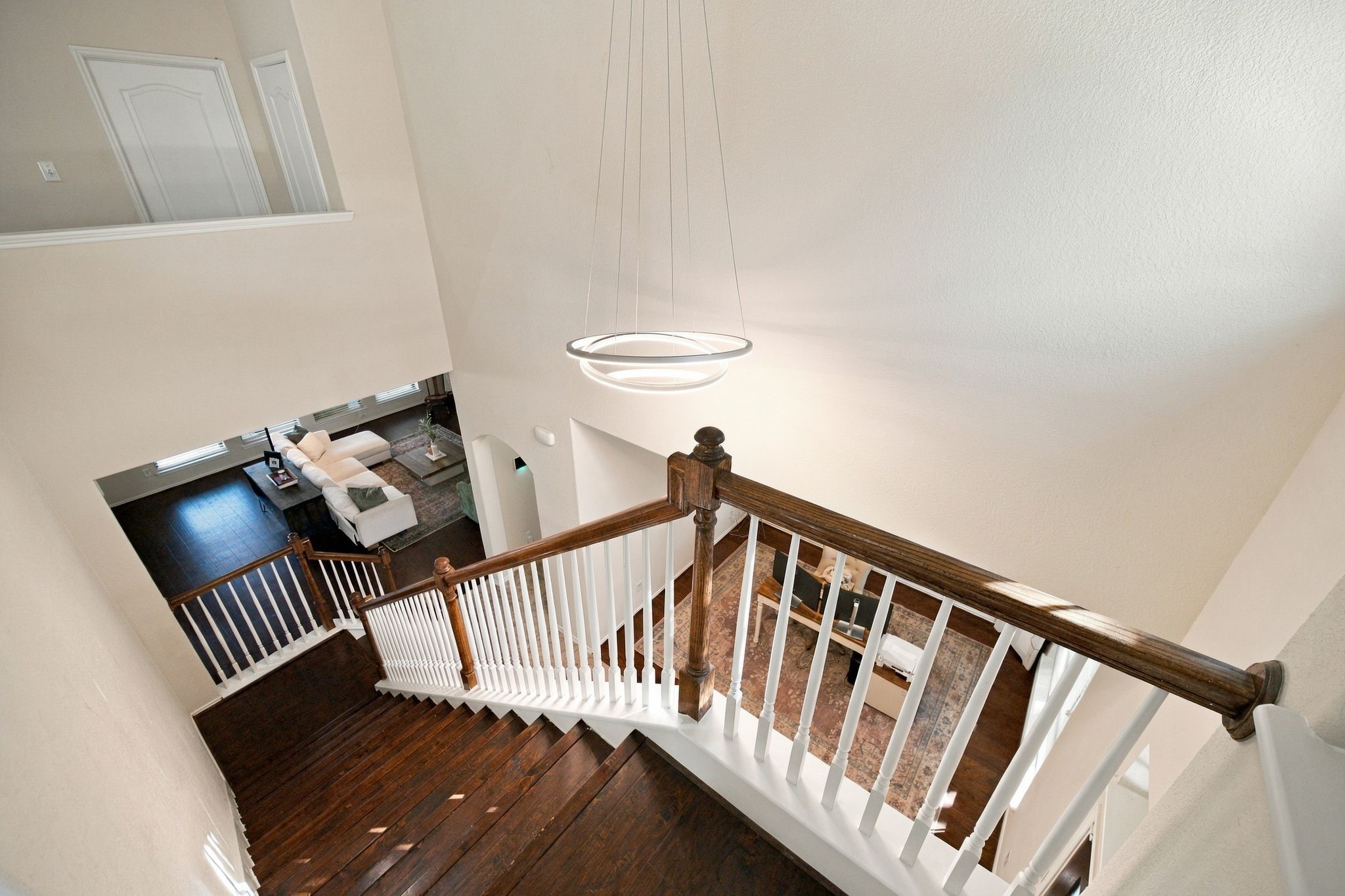 Elegant staircase with dark wood treads and white balusters leads to a bright, open living area featuring modern lighting and high ceilings.