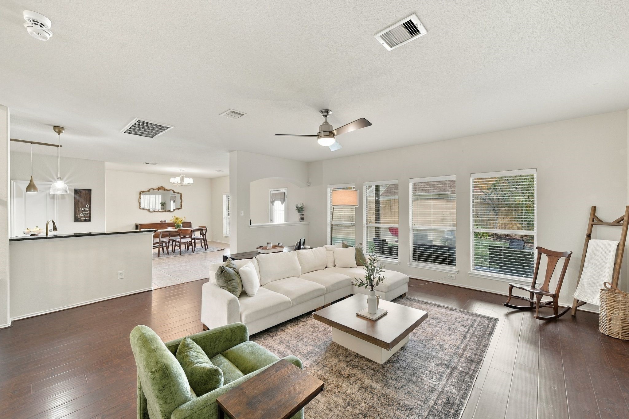 Spacious and bright open-concept living area with large windows, hardwood floors, and modern ceiling fan, seamlessly flowing into a dining space and kitchen with stylish pendant lighting.