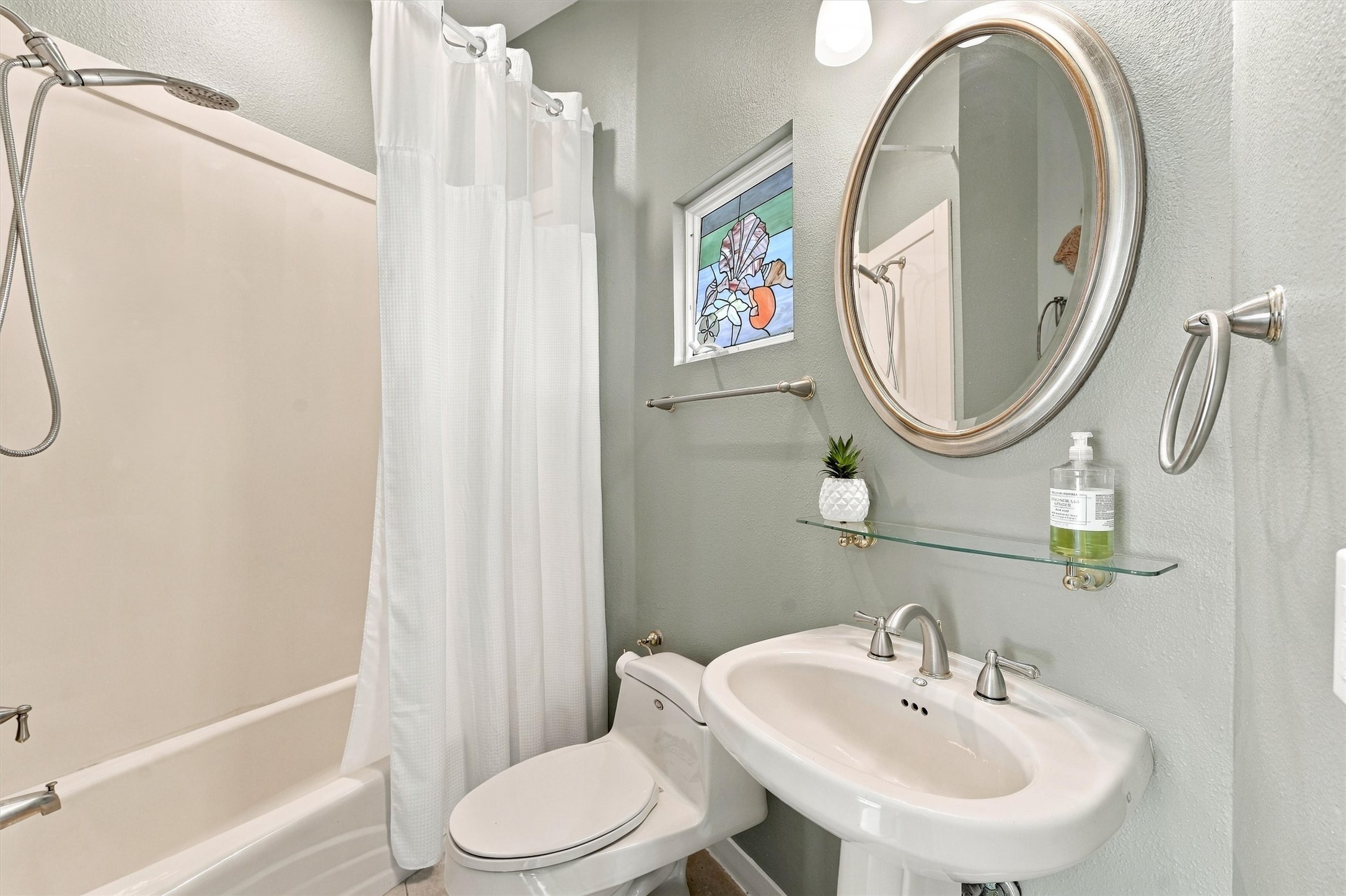 A colorful stained glass leans against the window adding seaside charm to this bath. A tub-and-shower combination, pedestal sink, and glass display shelf create a convenient place to freshen up.