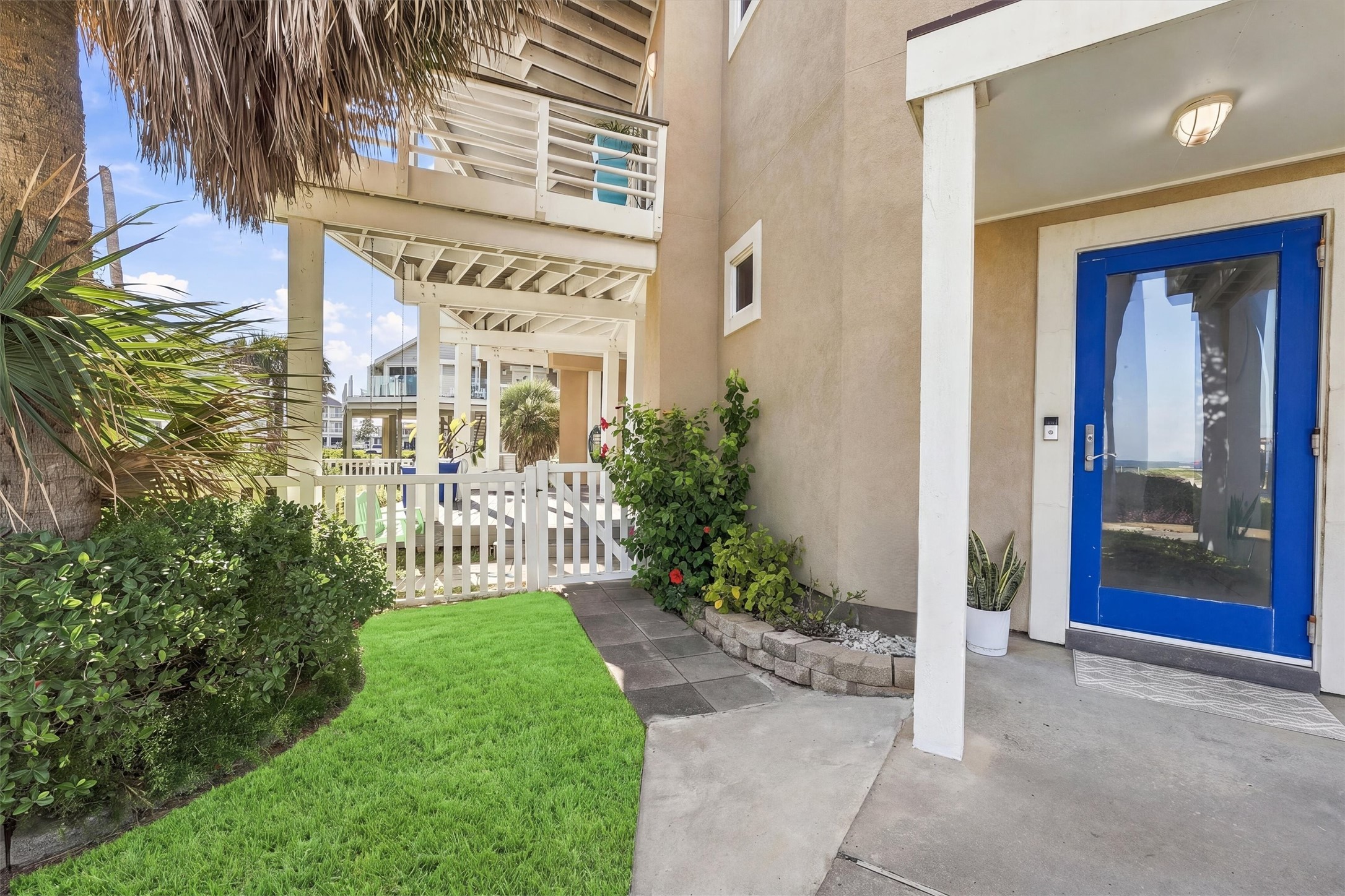 A vibrant blue door, tropical greenery, and a white picket gate welcome you home. This inviting entrance sets the tone for the relaxed island living.