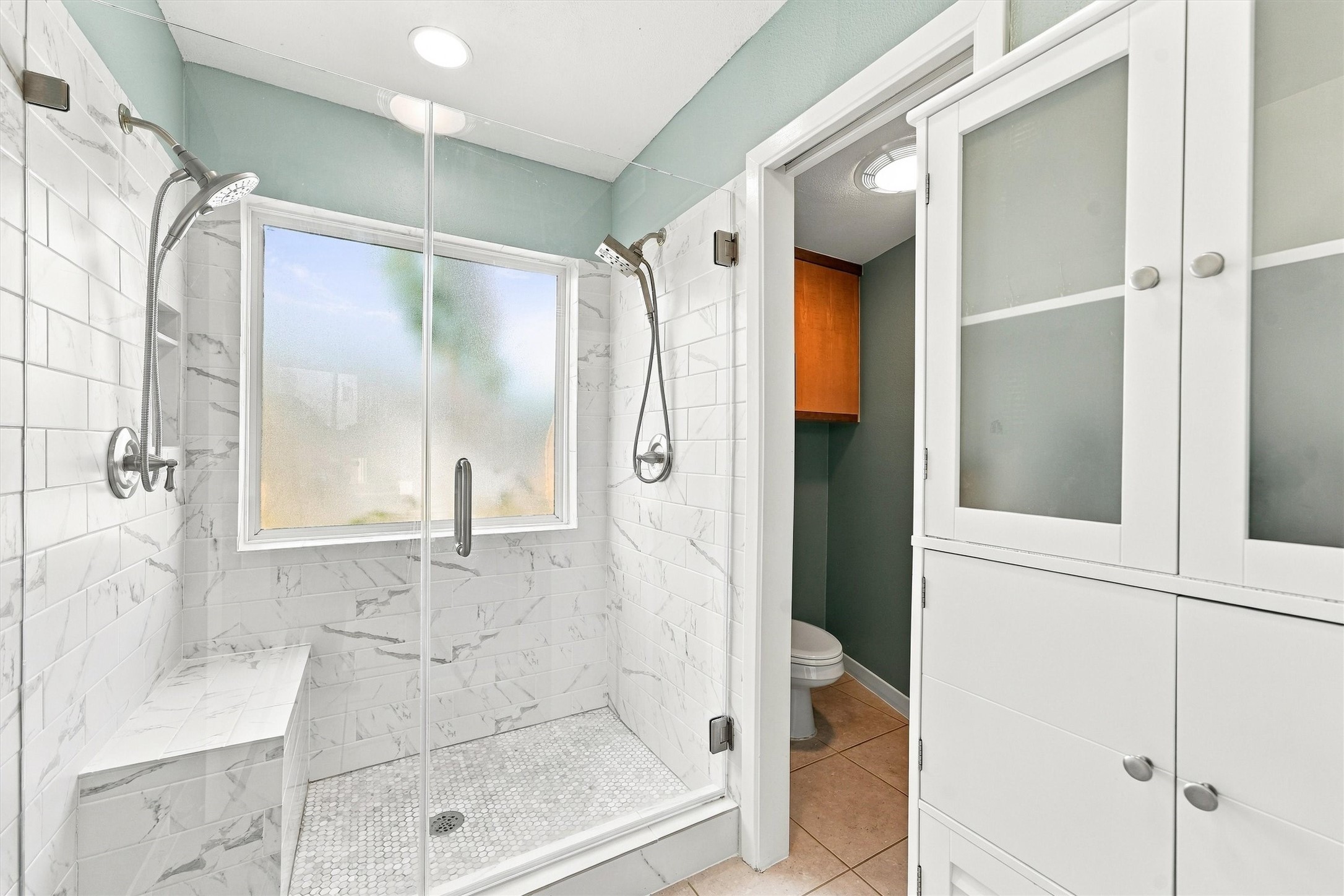A spacious glass-enclosed shower with dual showerheads and a built-in bench. Marble tile and soft natural light create a peaceful finish to your daily routine.