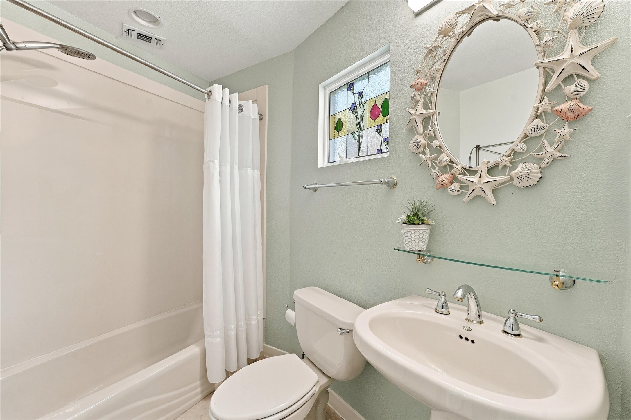 A tub-and-shower combination, pedestal sink, decorative stained glass resting against the window, bring convenience and seaside personality together.
