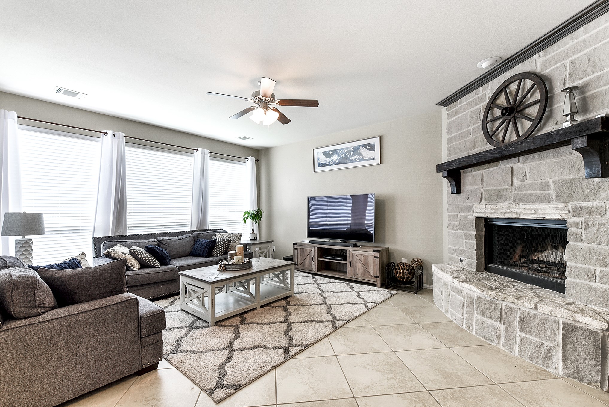 The living room features the same tile flooring, creating a cohesive flow throughout the home. A drop-down ceiling fan adds comfort and style, while a stone fireplace serves as a captivating focal point, enhancing the ambiance of the space.