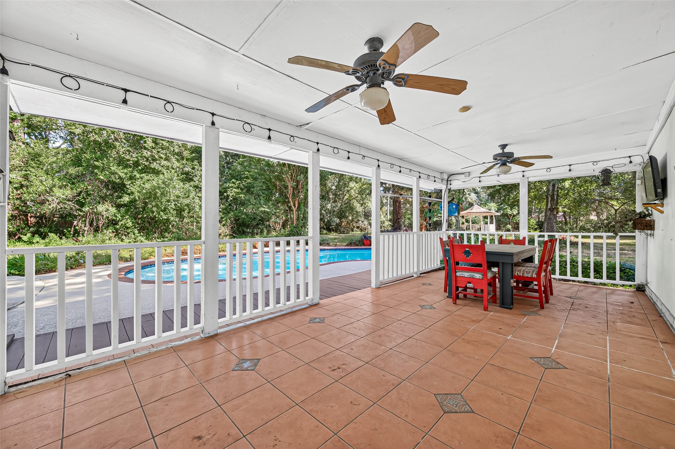 The covered patio has a terra cotta tile flooring, dual fans, outdoor tv, and surrounded with decorative lighting.
