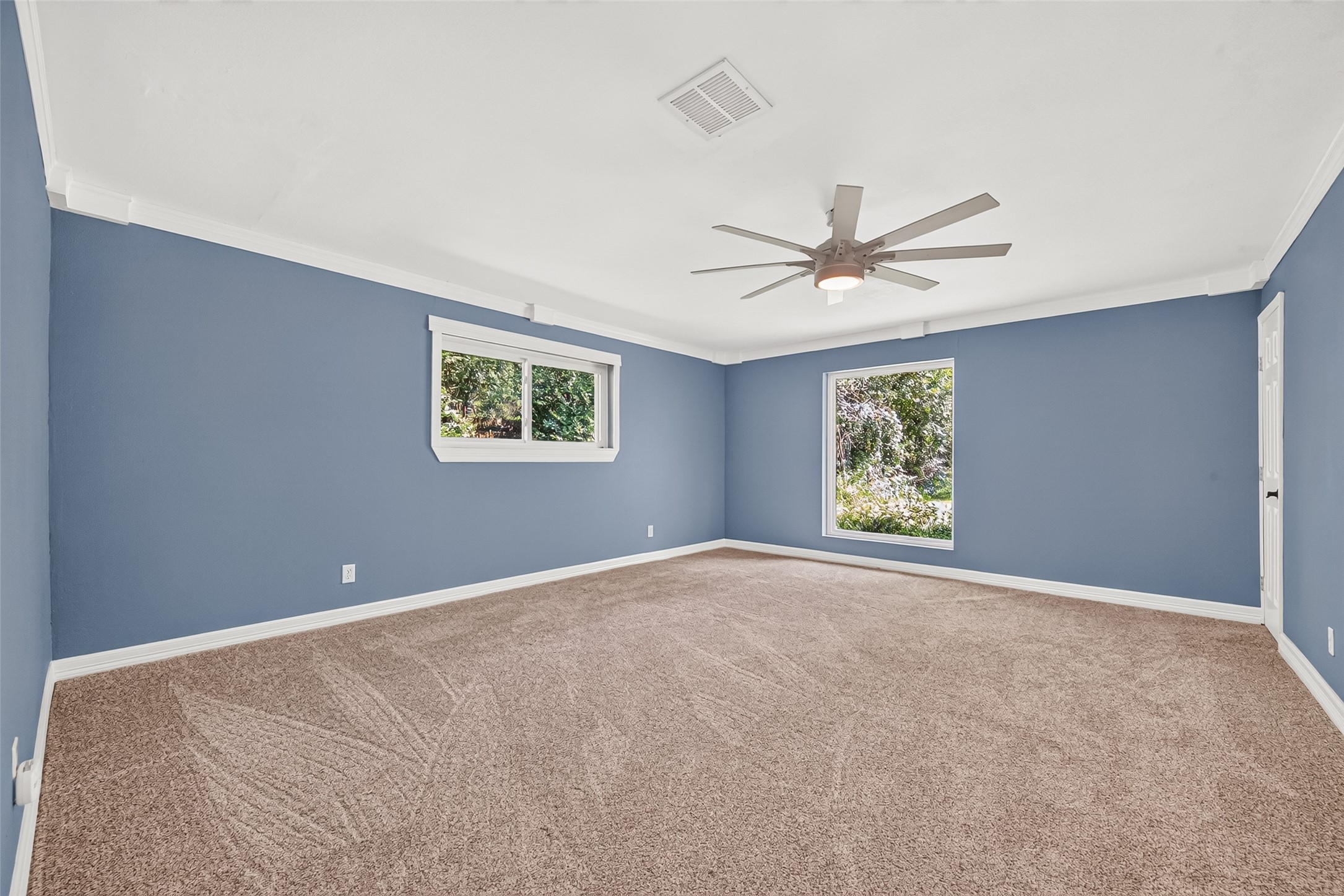 The spacious primary bedroom features ornate decorative crown molding, a large picture window overlooking the serene backyard, brand-new storm windows, plush carpeting, a modern seven-blade ceiling fan, and soothing new blue paint that creates a calm and inviting retreat.
